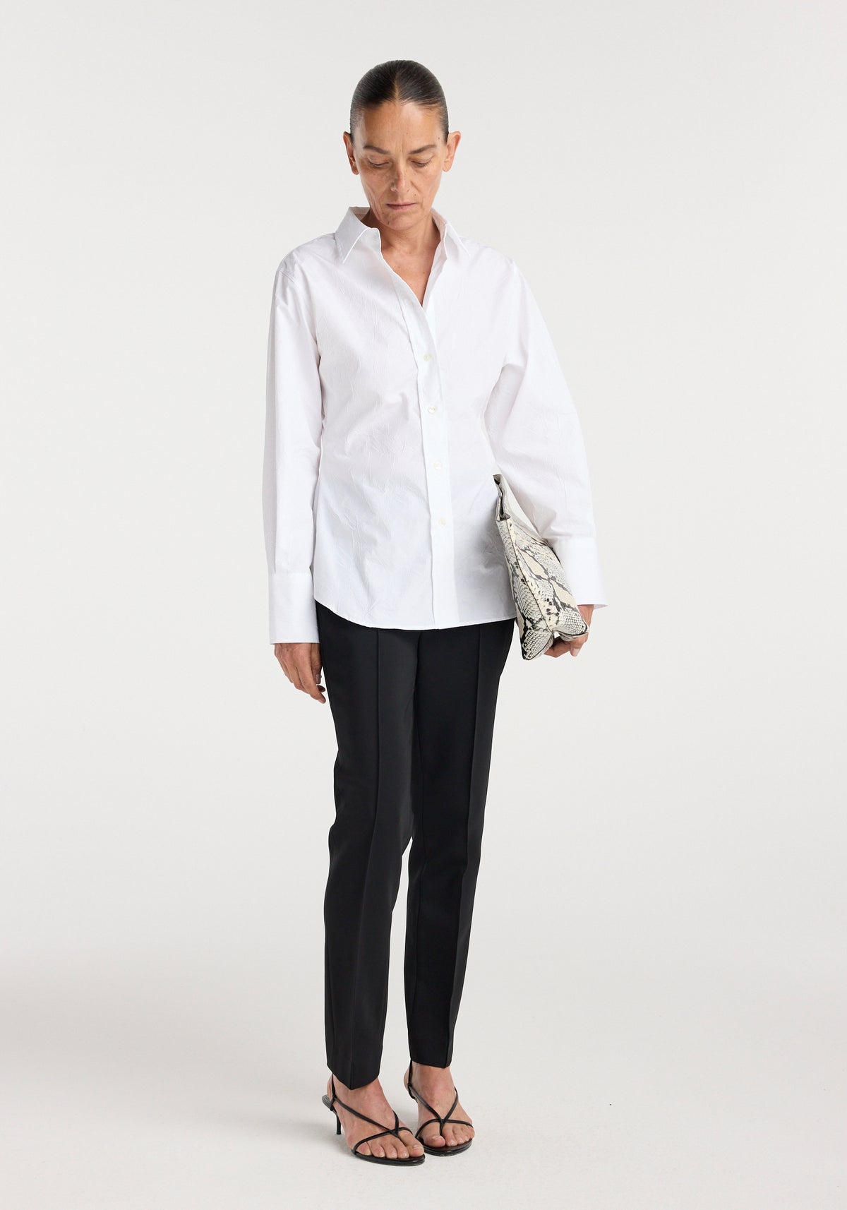 Woman wearing a shaped shirt | optic white paired with black tailored pants and strappy black heels, holding a snakeskin patterned clutch against a plain background.