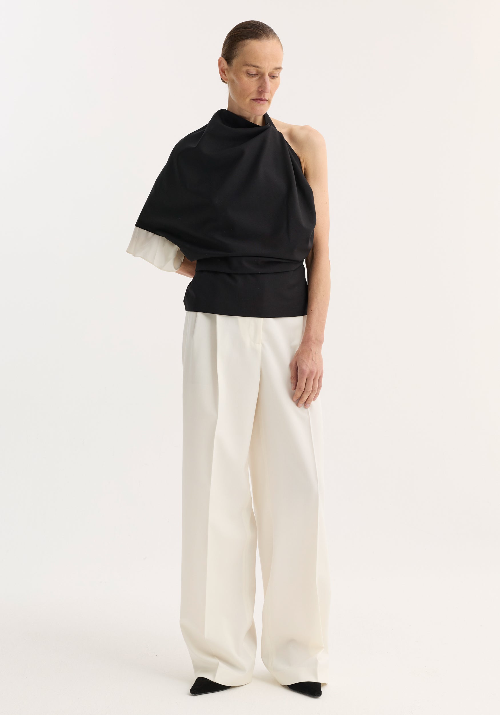 Woman wearing a Draped open back wool top | black paired with high-waisted wide-leg white pants and black pointed shoes, standing against a plain white background.