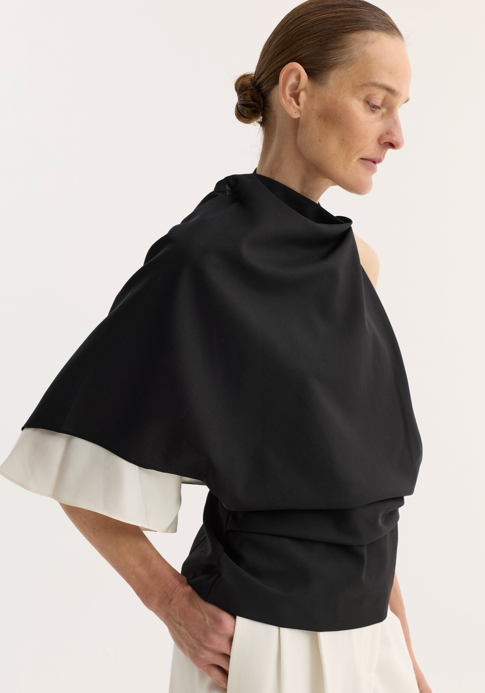Woman wearing a black Draped open back wool top | black over a white dress, shown in profile with hair tied back and looking downward.