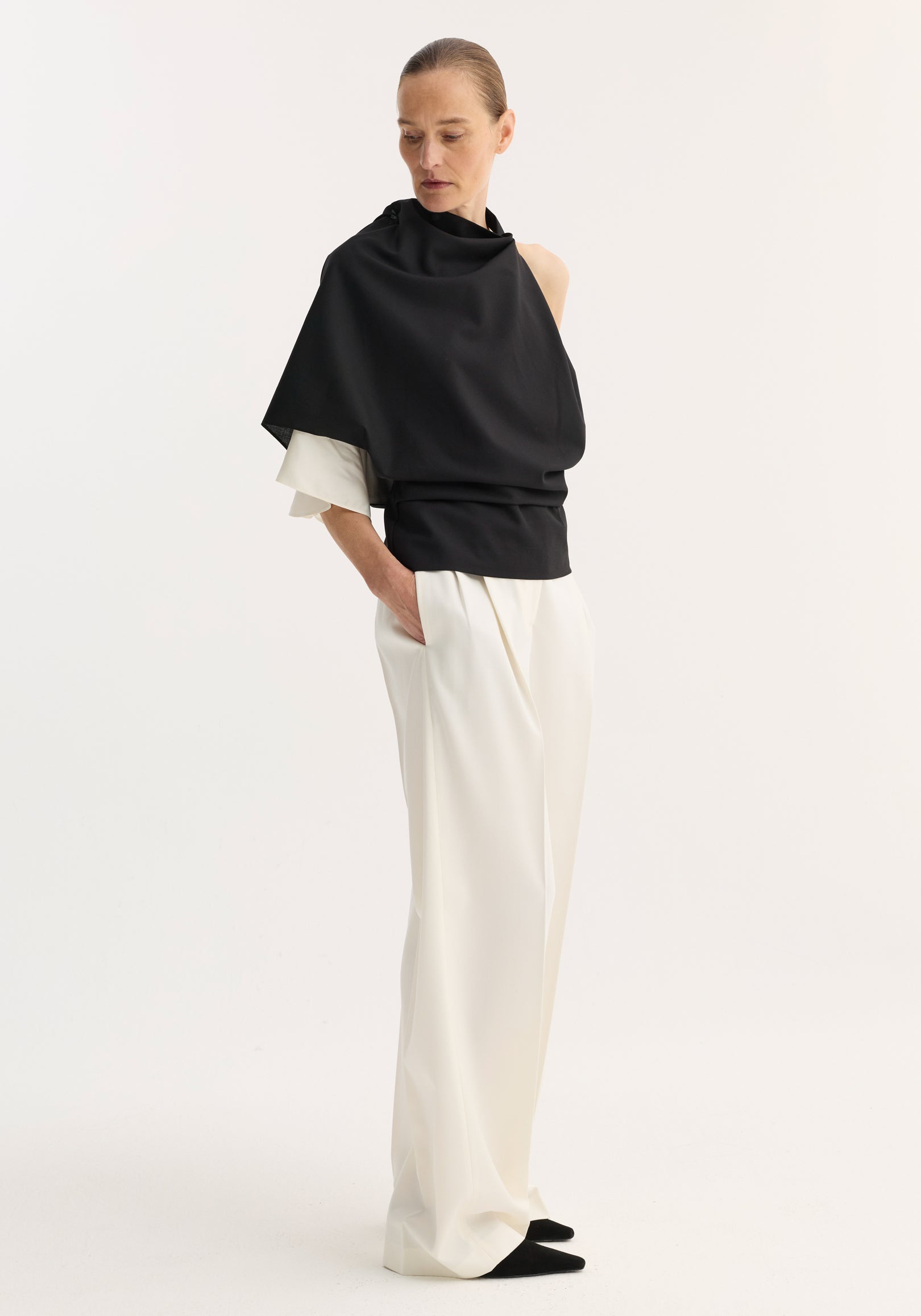 Woman wearing a black Draped open back wool top | black paired with loose white pants, standing with hands in pockets against a plain white background.