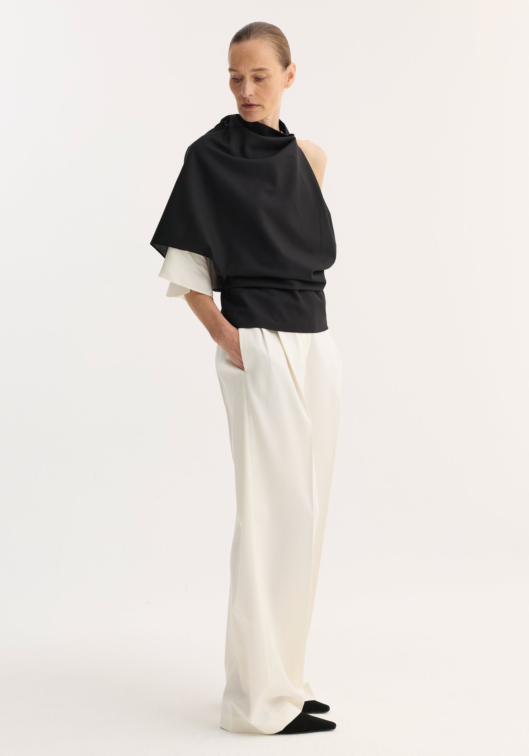 Woman wearing a black Draped open back wool top | black paired with loose white pants, standing with hands in pockets against a plain white background.