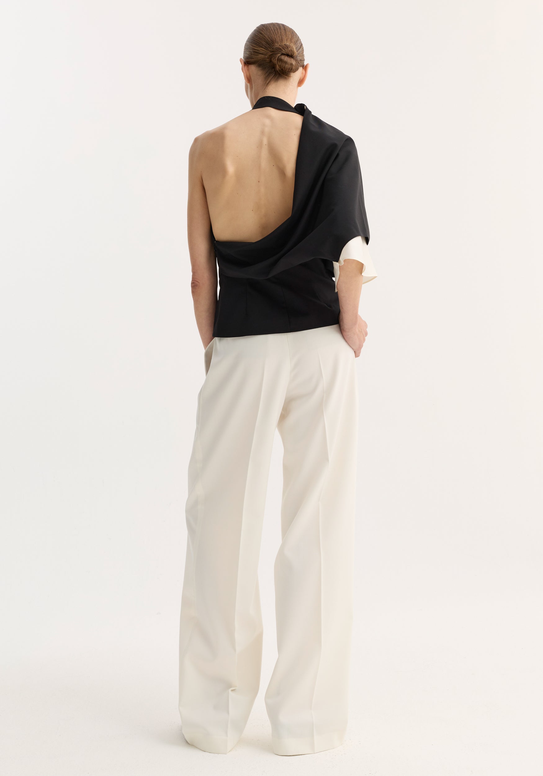 Model seen from the back wearing a Draped open back wool top | black with one sleeve draped over the shoulder, paired with loose white trousers against a plain background.