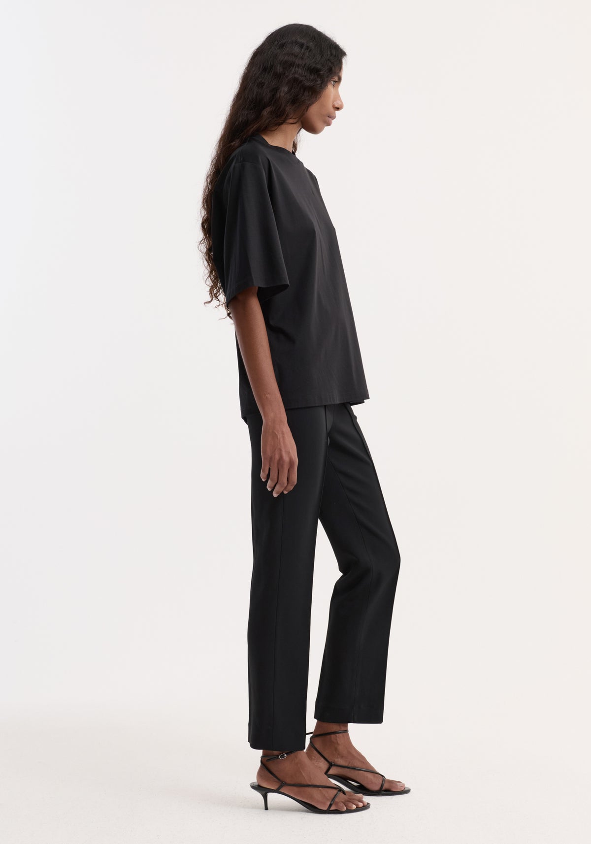 Side view of a model wearing the Oversized cotton silk t-shirt | black with matching black trousers and strappy heeled sandals.