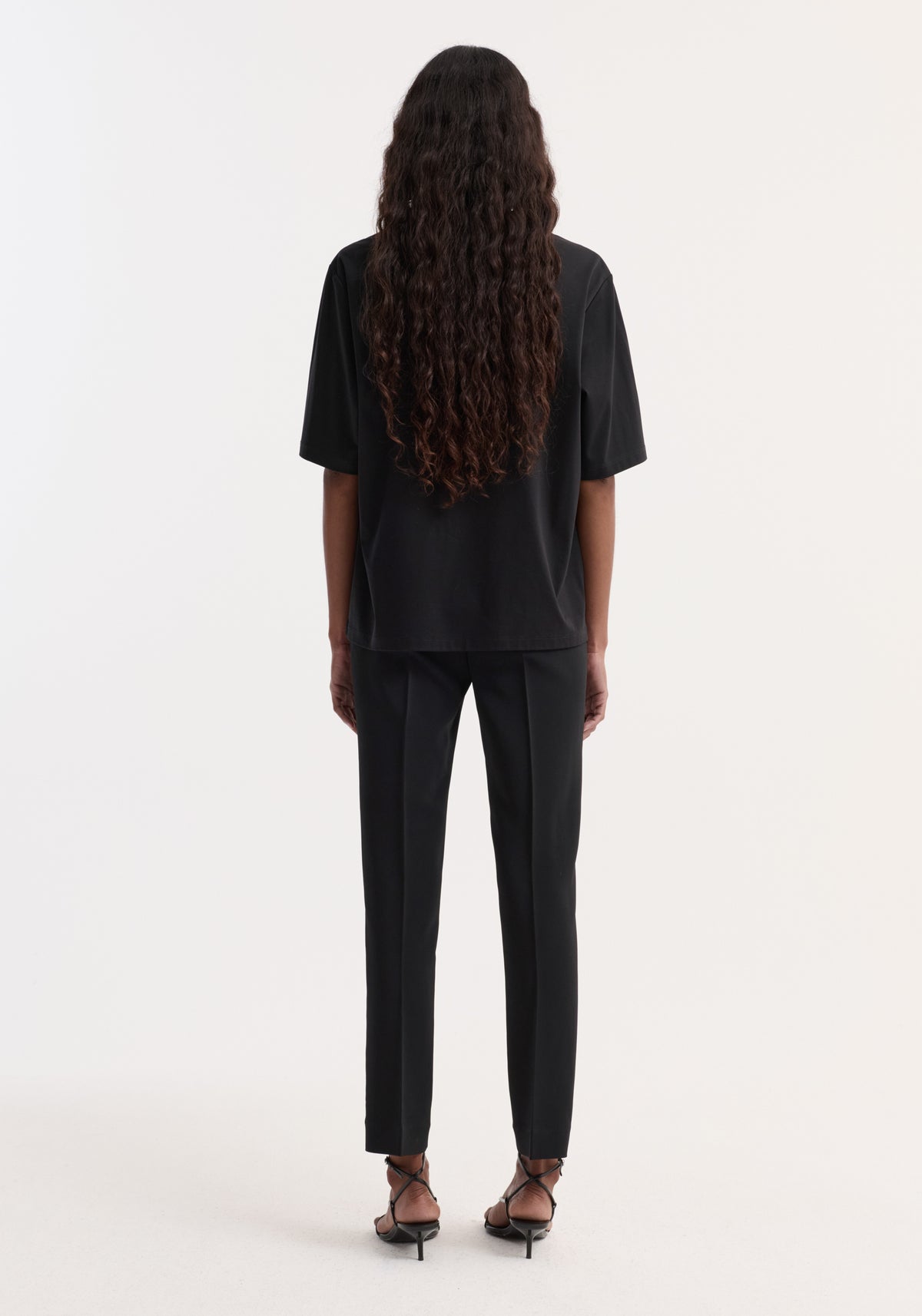 Back view of a model wearing Oversized cotton silk t-shirt | black with a loose fit and short sleeves, styled with black trousers and strappy heels