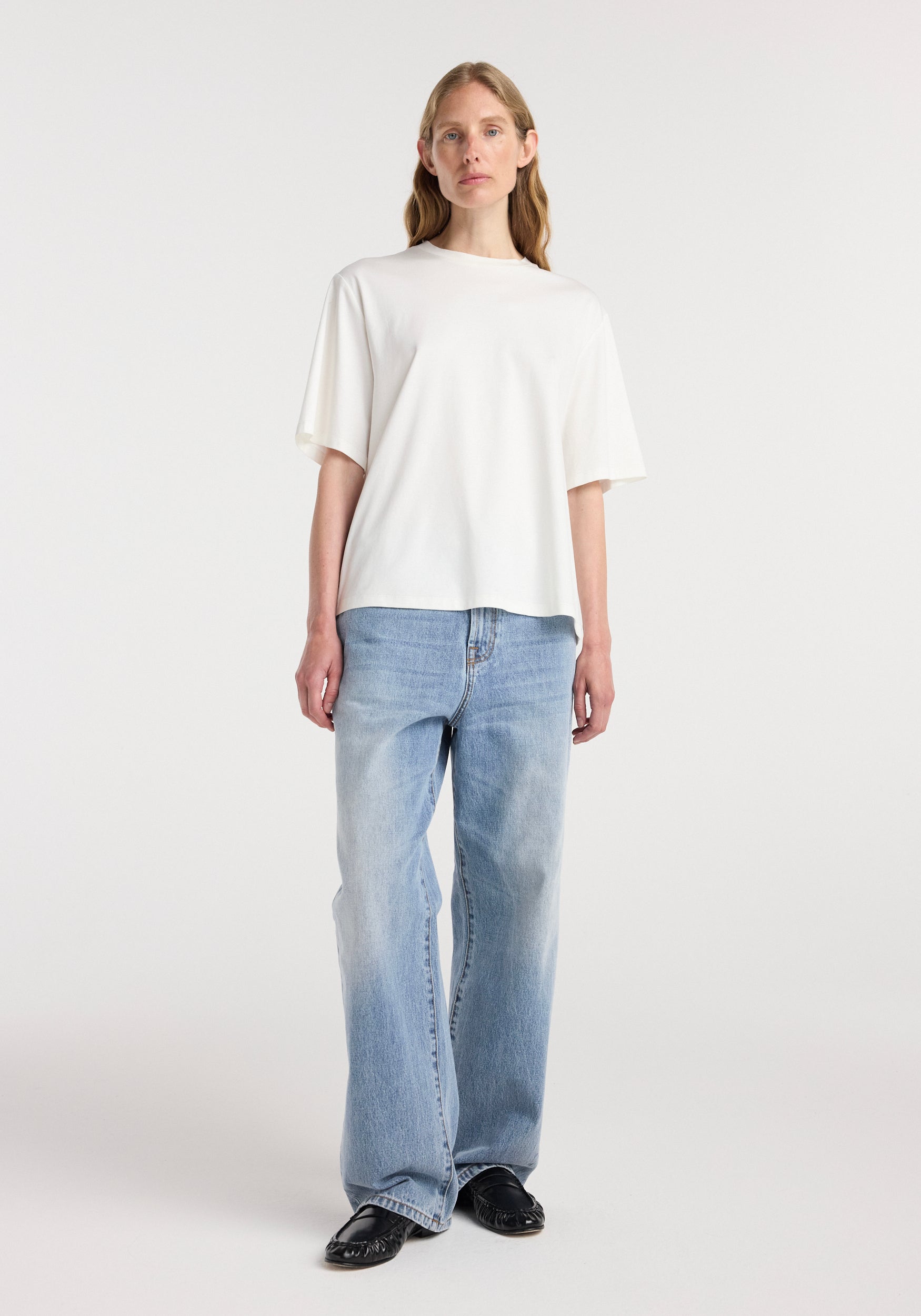 A woman wearing an oversized cotton silk t-shirt | off white paired with light blue wide-leg jeans and black shoes against a plain white background.