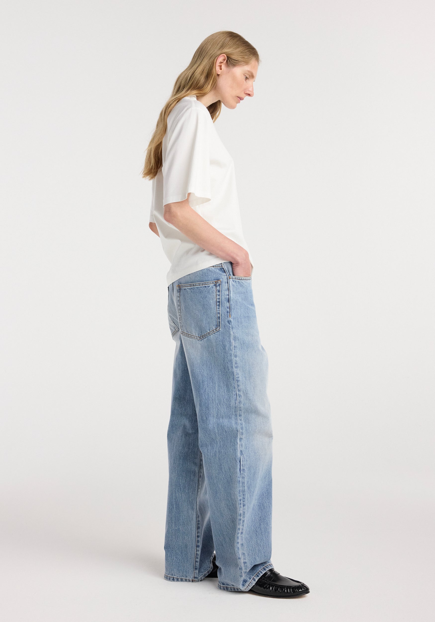 Profile view of a woman wearing an oversized cotton silk t-shirt | off white paired with light blue wide-leg jeans and black shoes against a plain background.