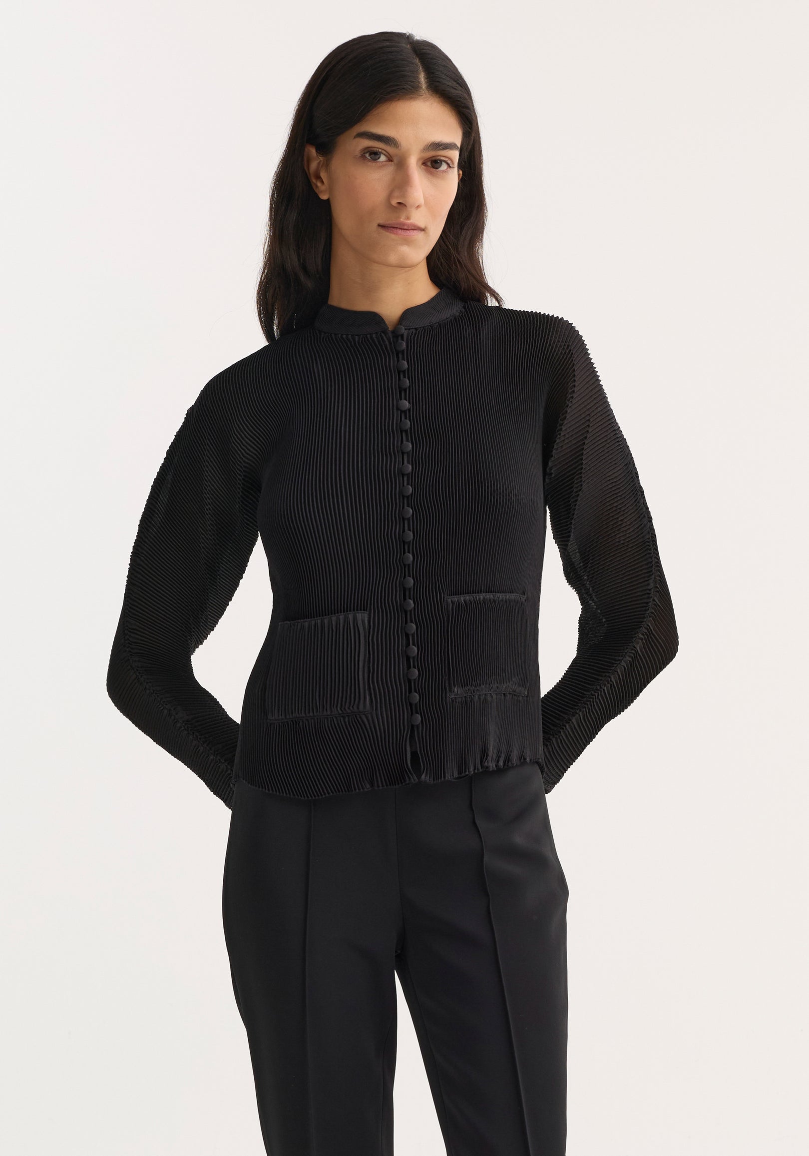 Woman wearing a black plissé cardigan | black with long sleeves, button-down front, and front pockets, paired with black tailored pants against a plain white background.