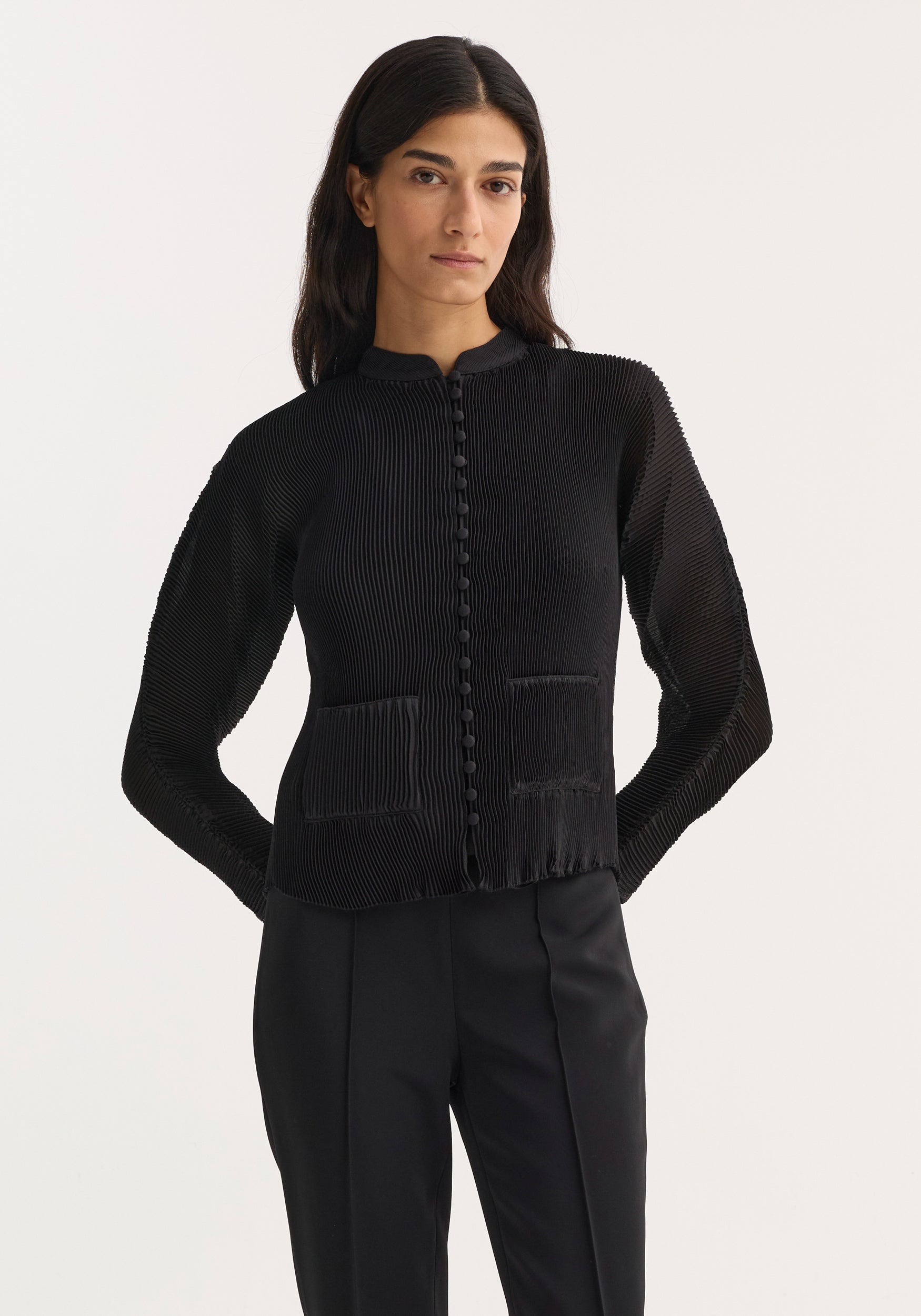 Woman wearing a black plissé cardigan | black with long sleeves, button-down front, and front pockets, paired with black tailored pants against a plain white background.