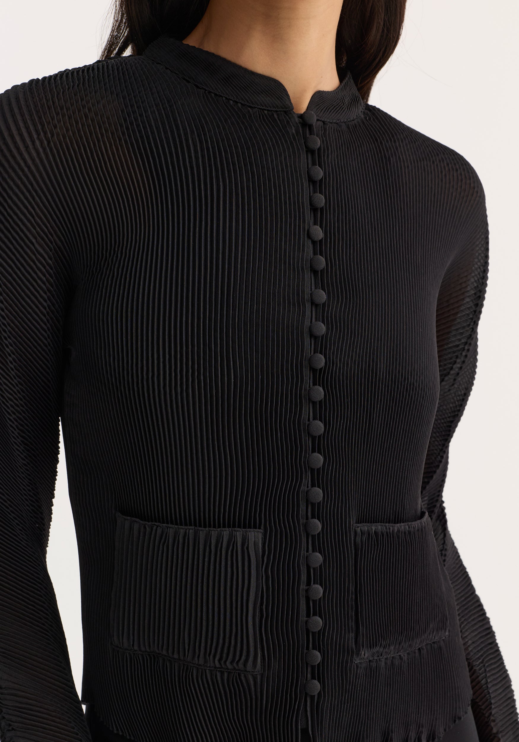 Close-up of a person wearing a black Plissé cardigan | black with vertical pleats, a row of fabric-covered buttons down the front, and two front pockets.