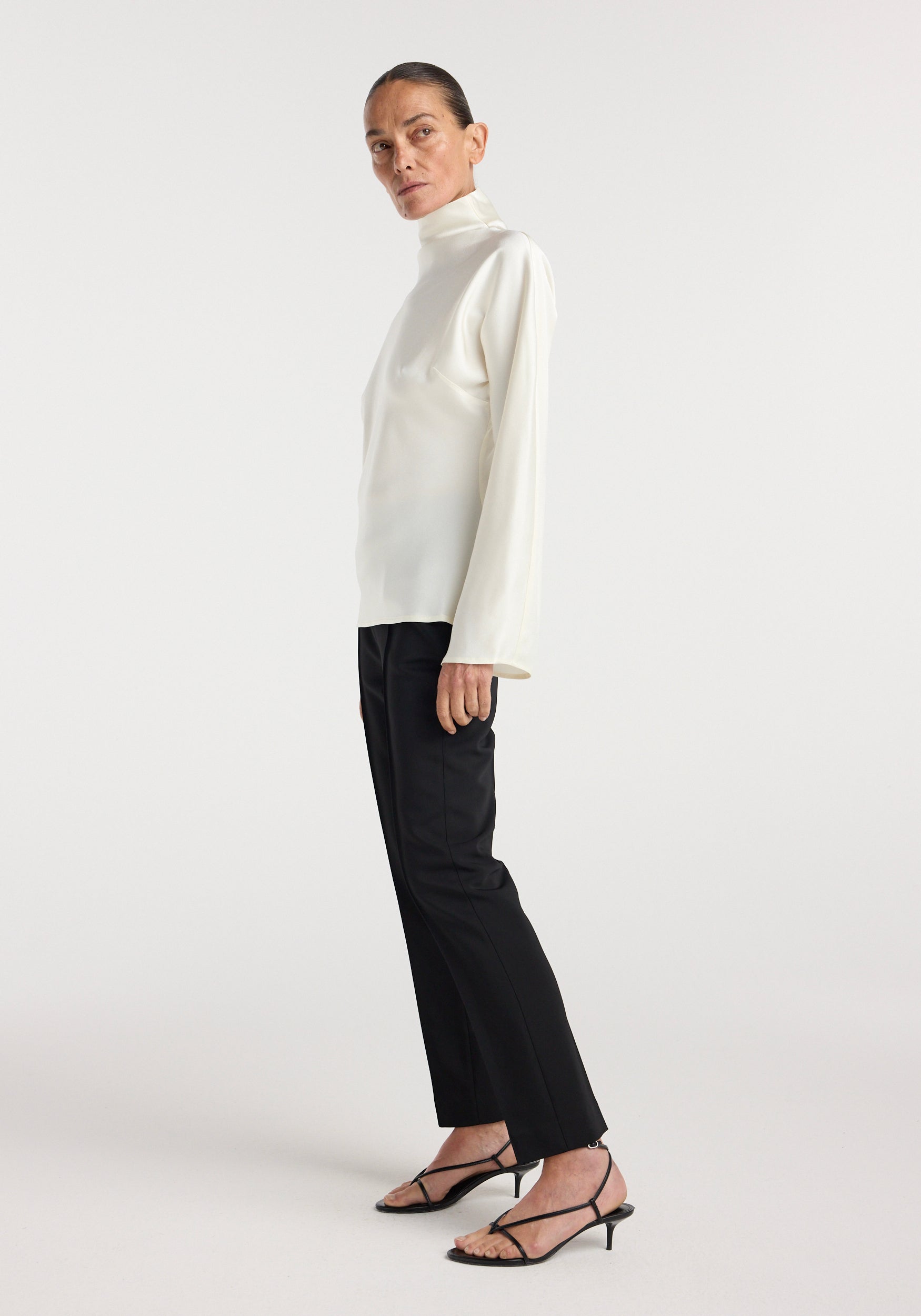 Woman wearing a cream satin waisted top featuring a high neck and long sleeves, paired with black tailored pants and black strappy heels.