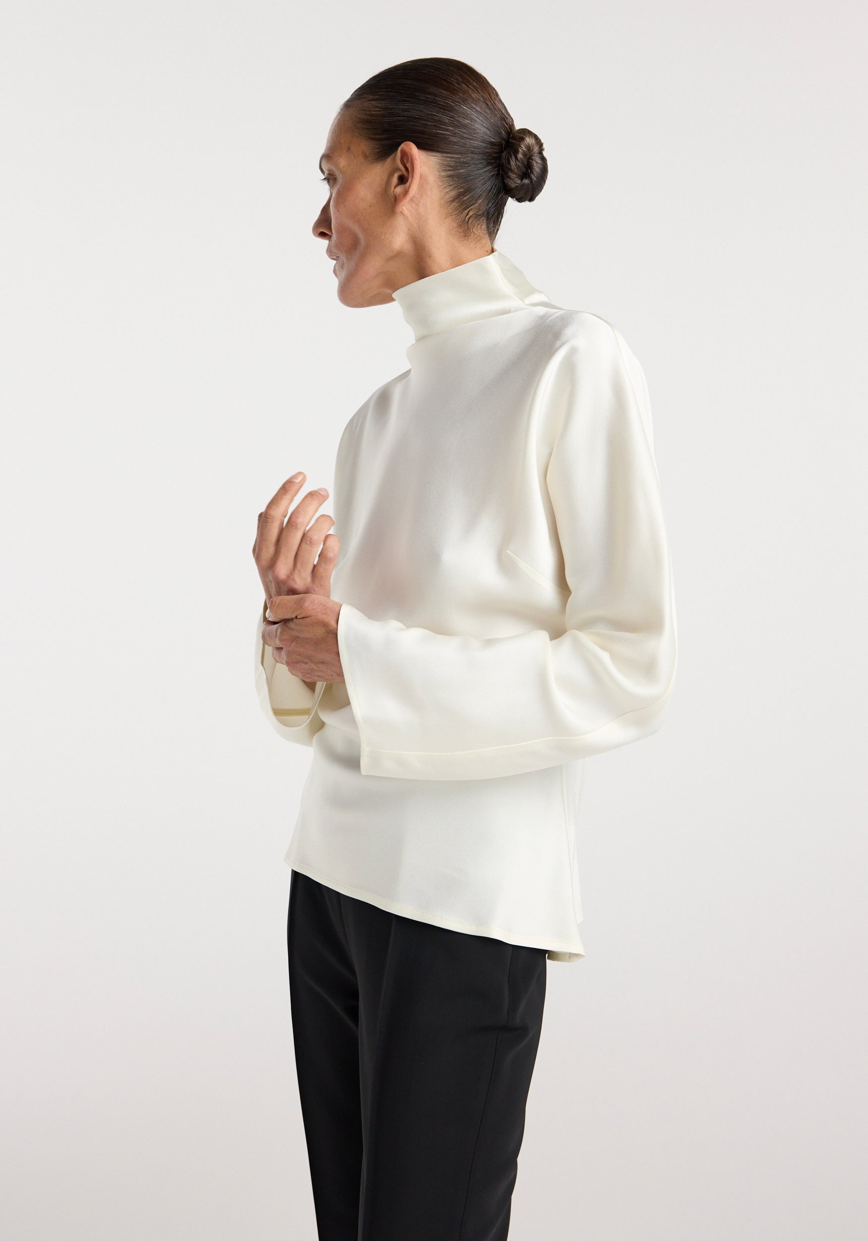 Woman wearing a cream high-neck Satin waisted top with long sleeves, paired with black pants, photographed against a plain light background.