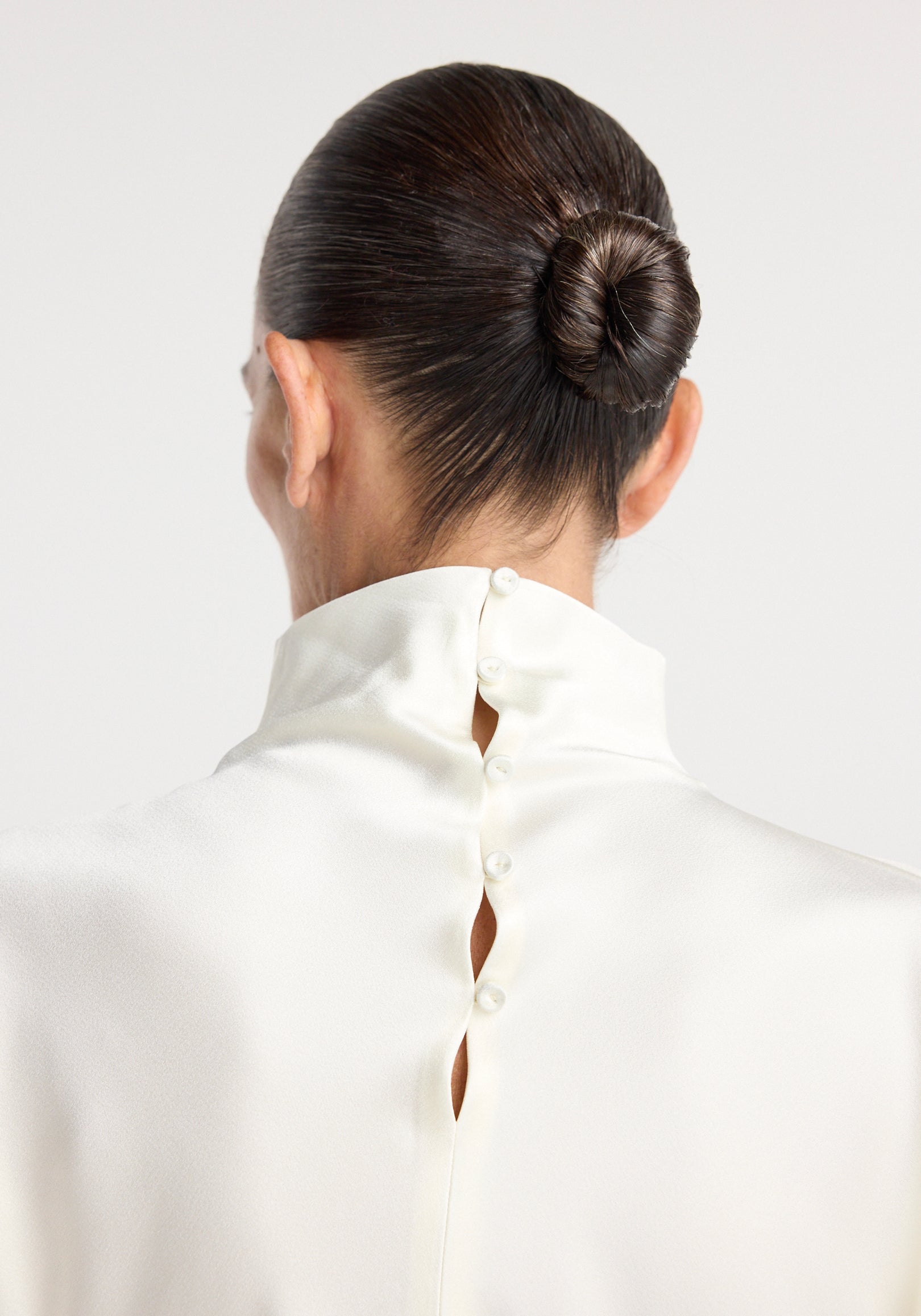 Close-up rear view of a person wearing a cream satin waisted top with a high collar and button details, hair styled in a sleek bun.
