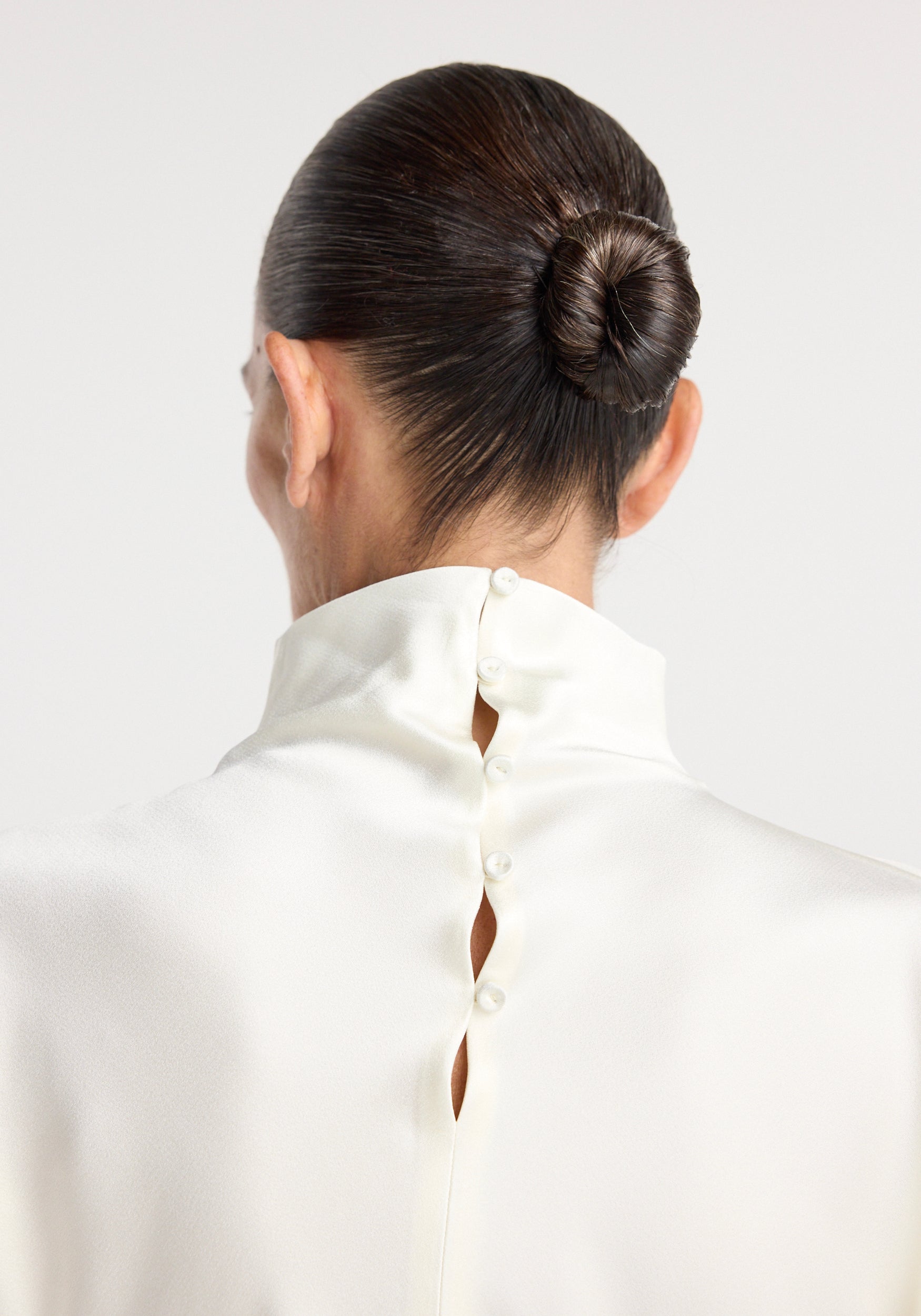 Close-up rear view of a person wearing a cream satin waisted top with a high collar and button details, hair styled in a sleek bun.