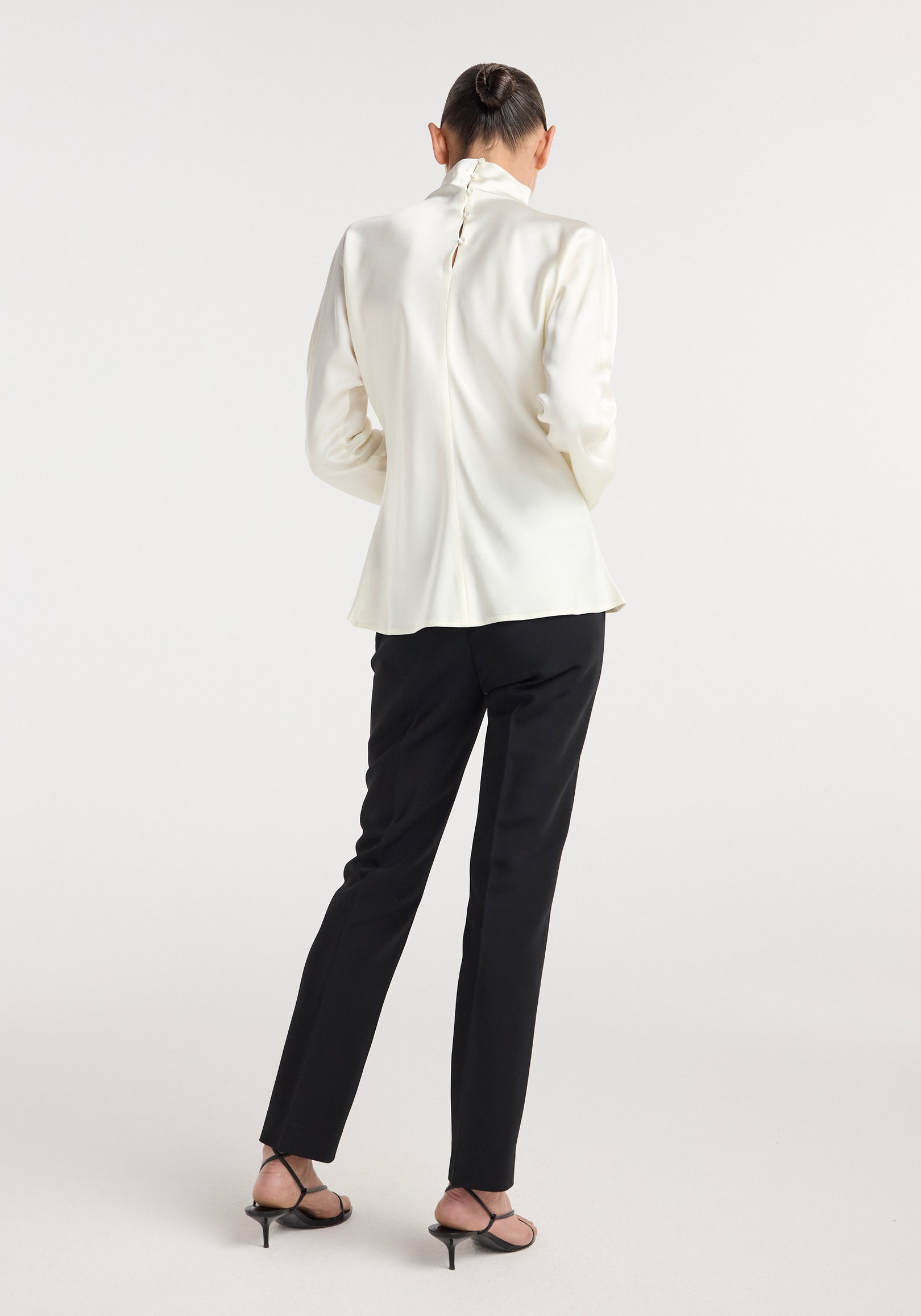 Woman wearing a cream-colored Satin waisted top with long sleeves and a high collar, paired with black slim pants and black strappy heels, posing with her back to the camera.