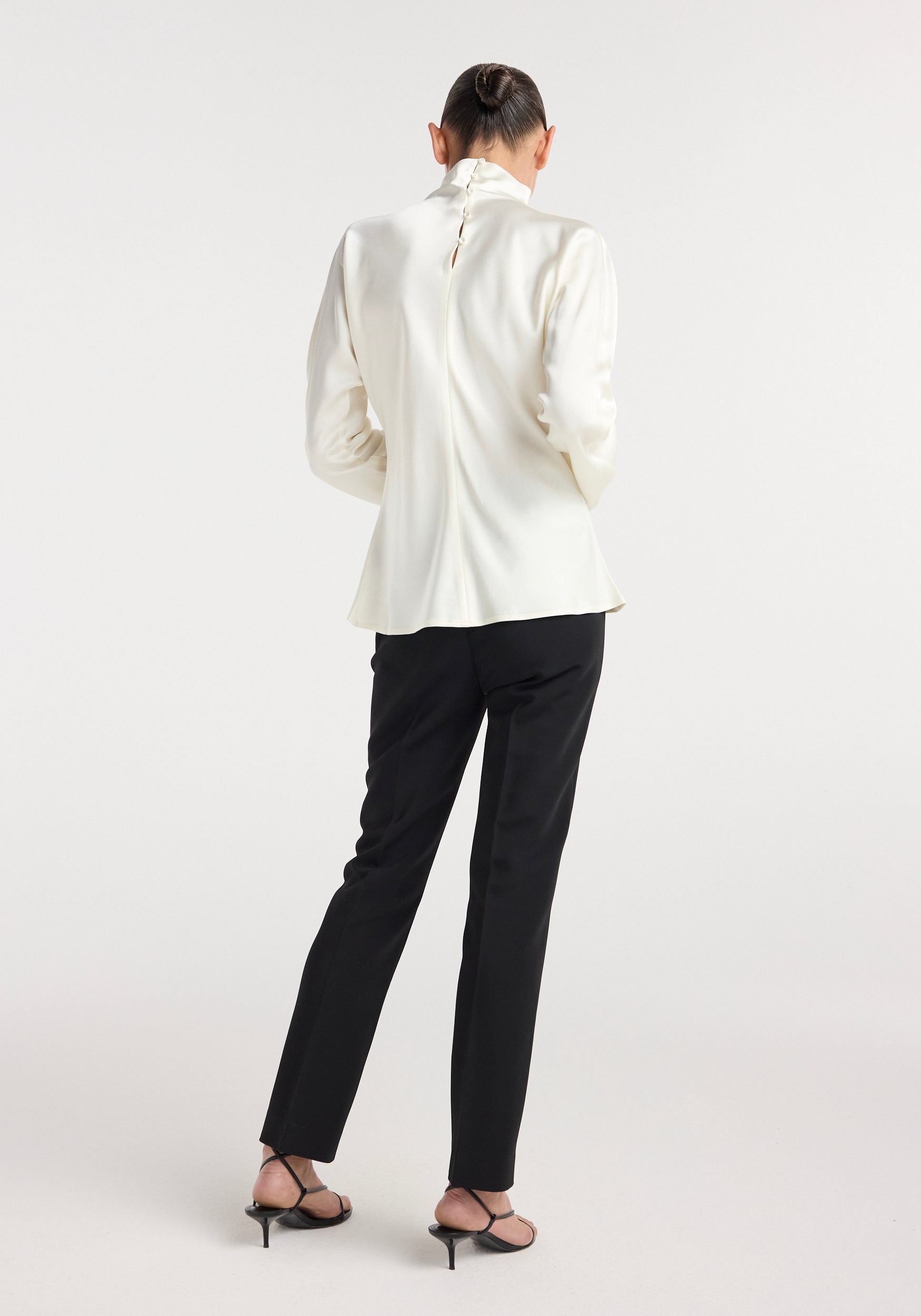 Woman wearing a cream-colored Satin waisted top with long sleeves and a high collar, paired with black slim pants and black strappy heels, posing with her back to the camera.