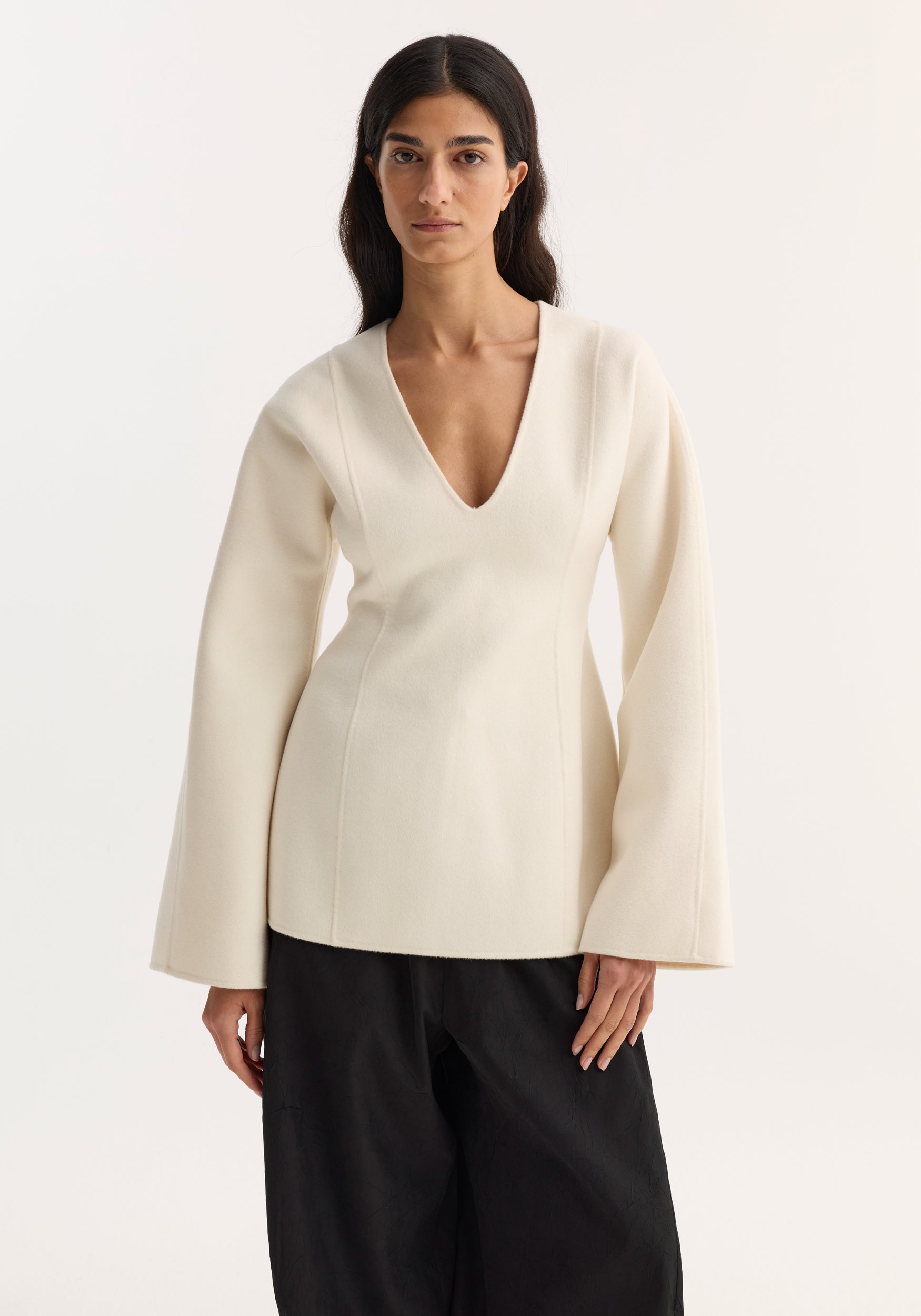 Woman wearing a sculptural long sleeve top | off white with a deep V neckline and flared sleeves paired with black wide-leg pants against a plain background.