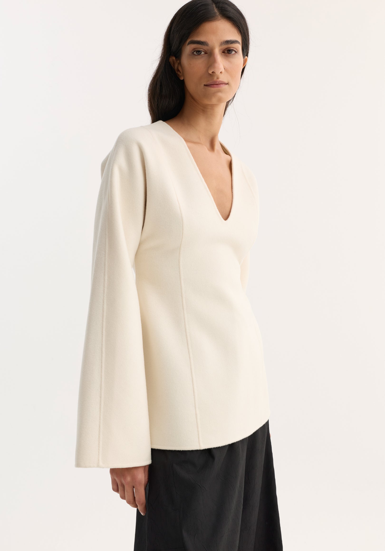 Woman wearing an off white sculptural long sleeve top with a deep V neckline and wide sleeves, paired with black pants against a neutral background.