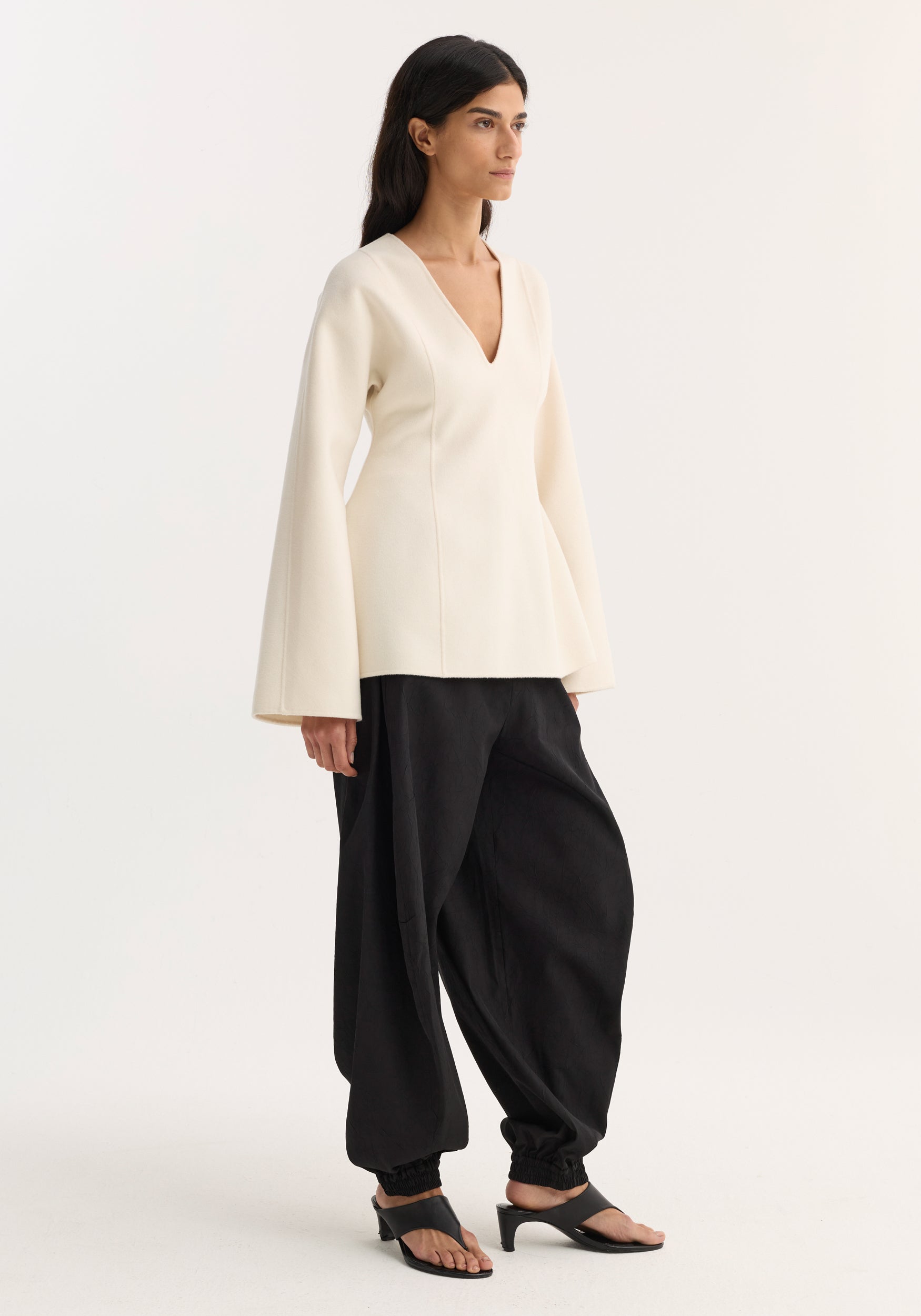 Woman wearing a sculptural long sleeve top | off white with a deep V-neck and flared sleeves, paired with loose black pants and open-toe black heeled sandals.