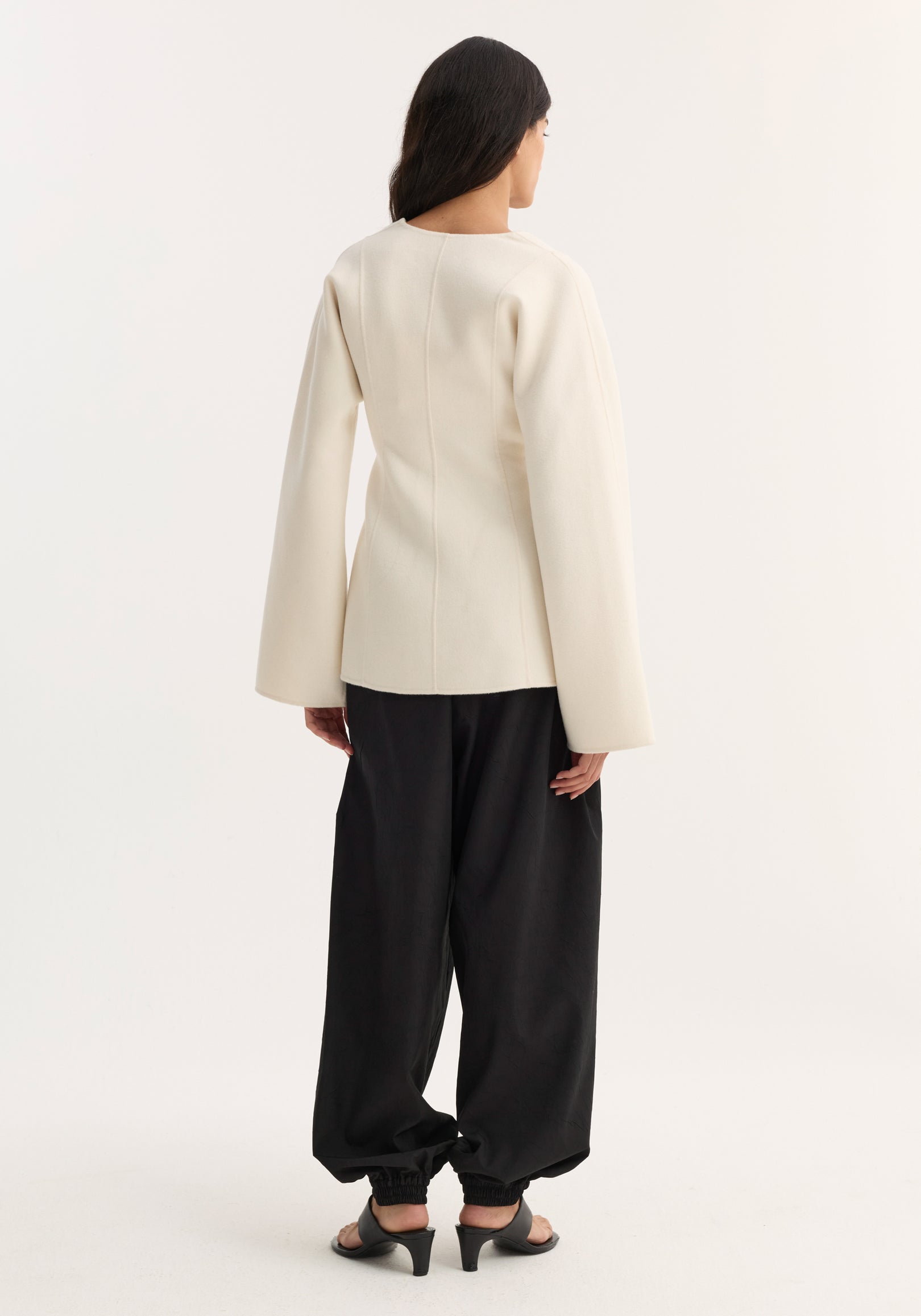 Woman seen from behind wearing a Sculptural long sleeve top | off white with a fitted back and slightly flared sleeves, paired with loose black pants and black heeled sandals.