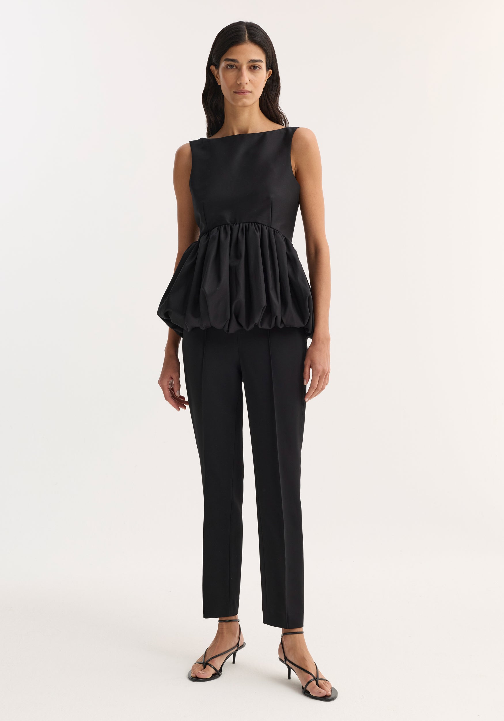 Woman wearing a sleeveless Voluminous boat-neck top | black with a gathered balloon hem paired with slim black trousers and black strappy heels against a plain background.