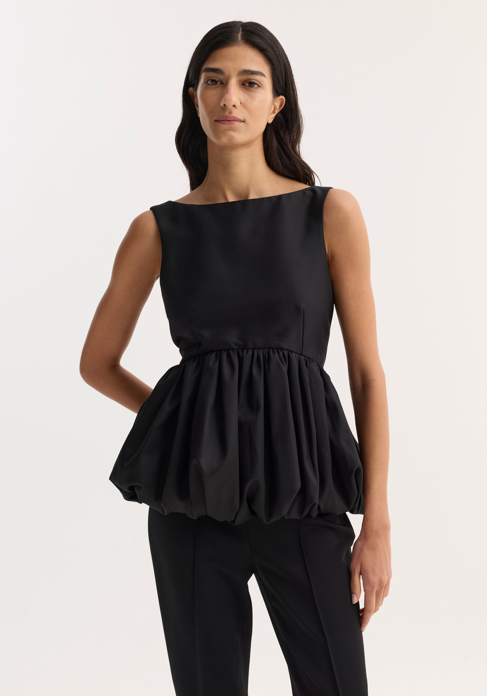 Woman wearing a voluminous boat-neck top | black with a gathered, puffed waist detail paired with sleek black pants against a plain light background.