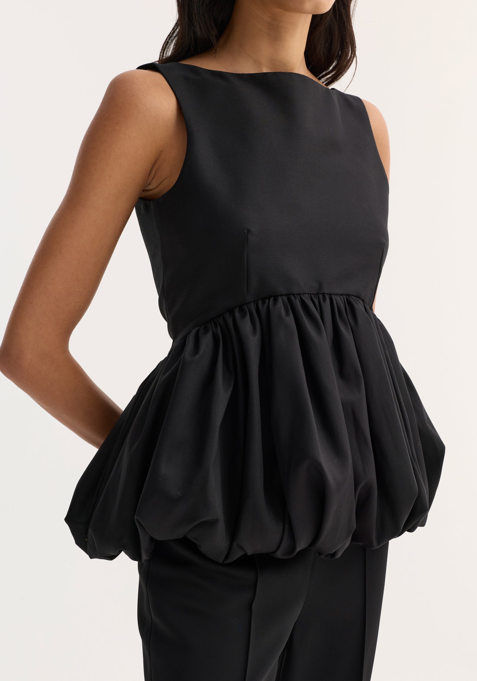 Woman wearing a black voluminous boat-neck top | black with a gathered, balloon-style hem and sleeveless design.