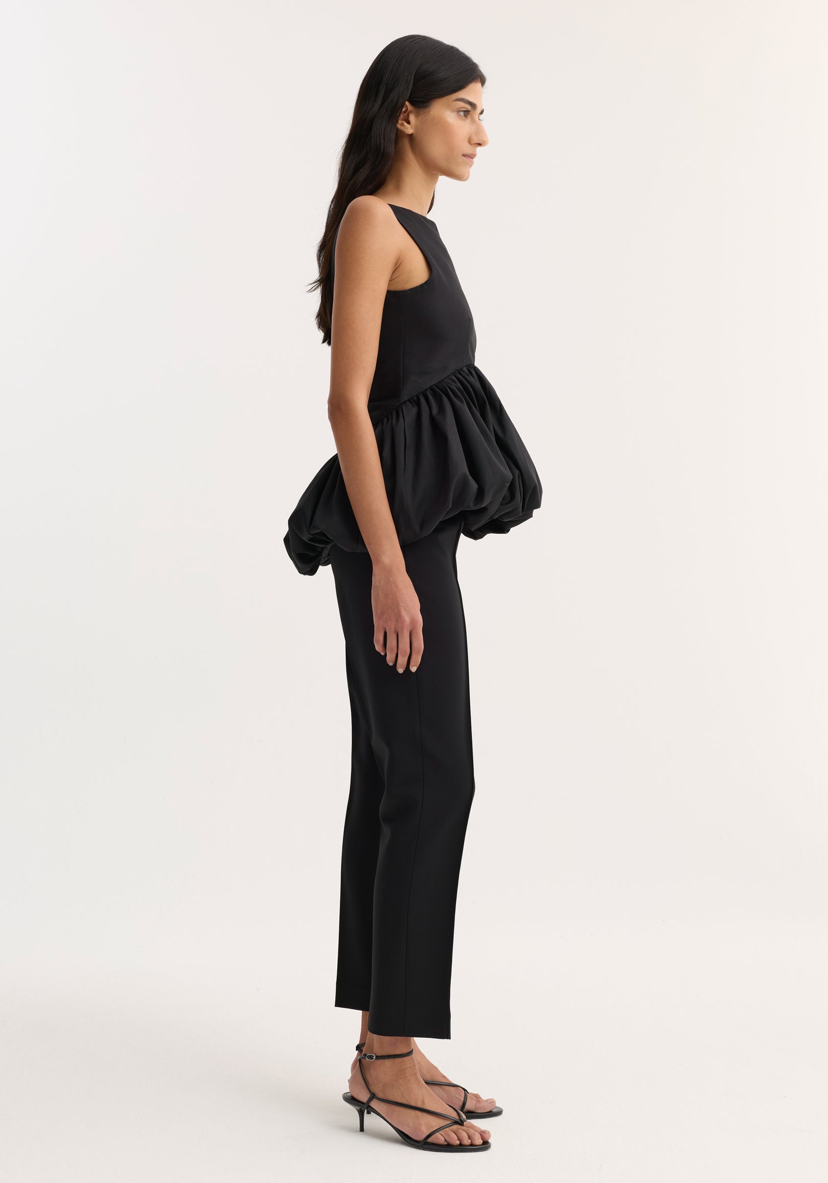 Side view of a woman wearing a black Voluminous boat-neck top | black with a puffed, gathered waist paired with black straight-leg pants and strappy black heels against a plain background.