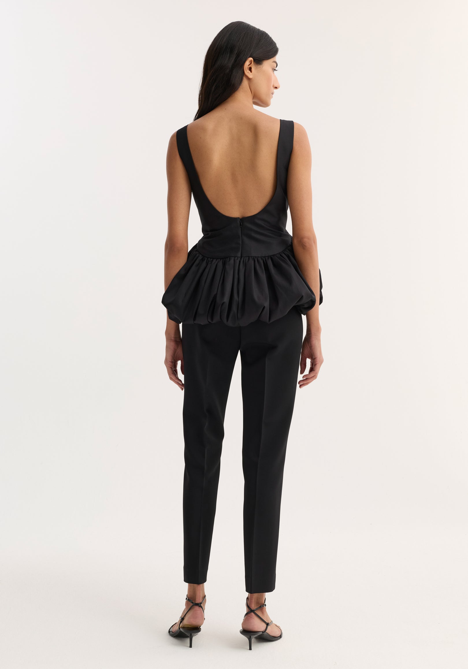 Woman wearing a voluminous boat-neck top | black with an open low back and gathered peplum detail, paired with slim black trousers and strappy black heels, standing facing away.