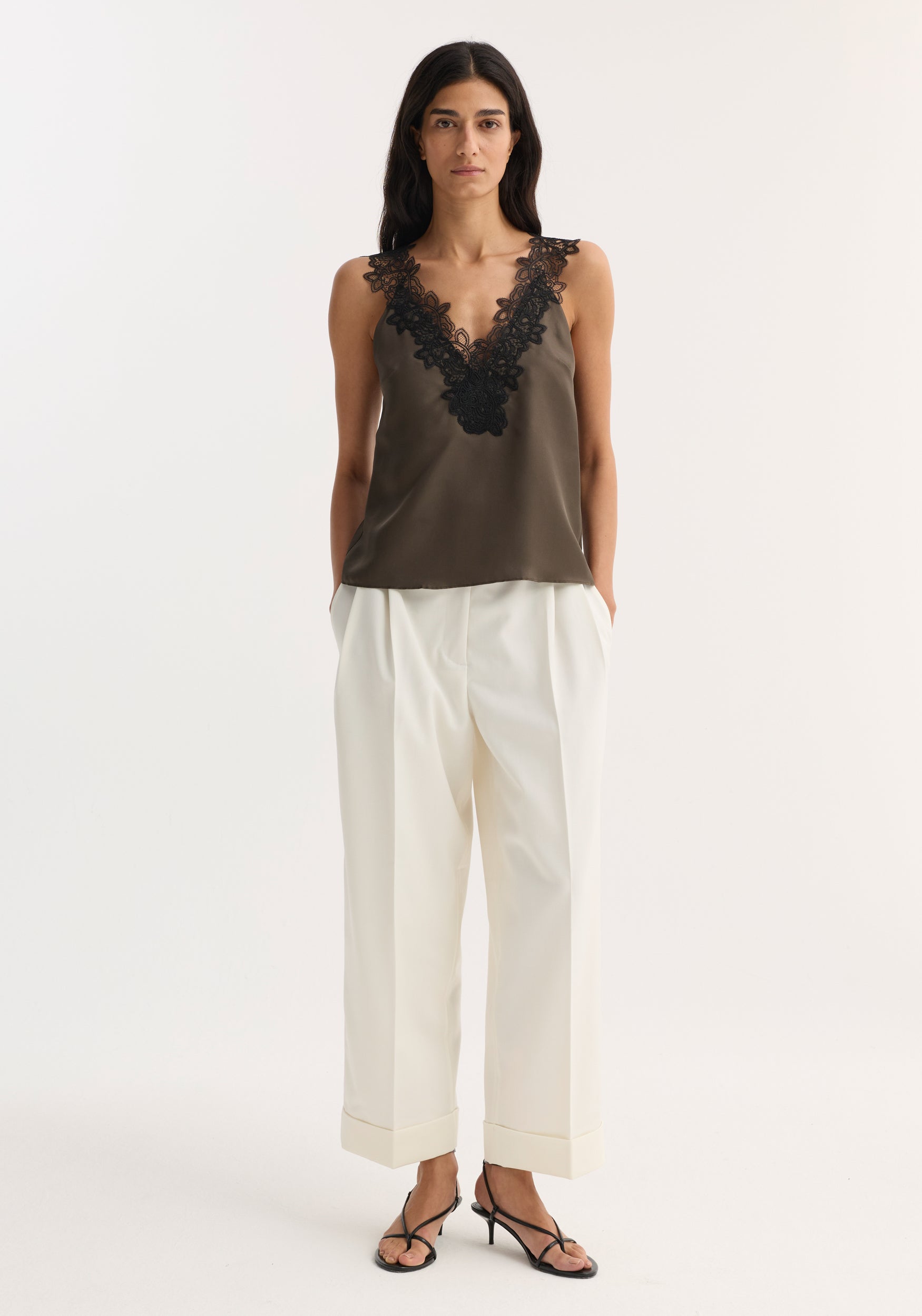 Woman wearing a sleeveless V-neck silk lace top in earth color with black lace details, paired with high-waisted white pants and black strappy heels, standing against a plain background.
