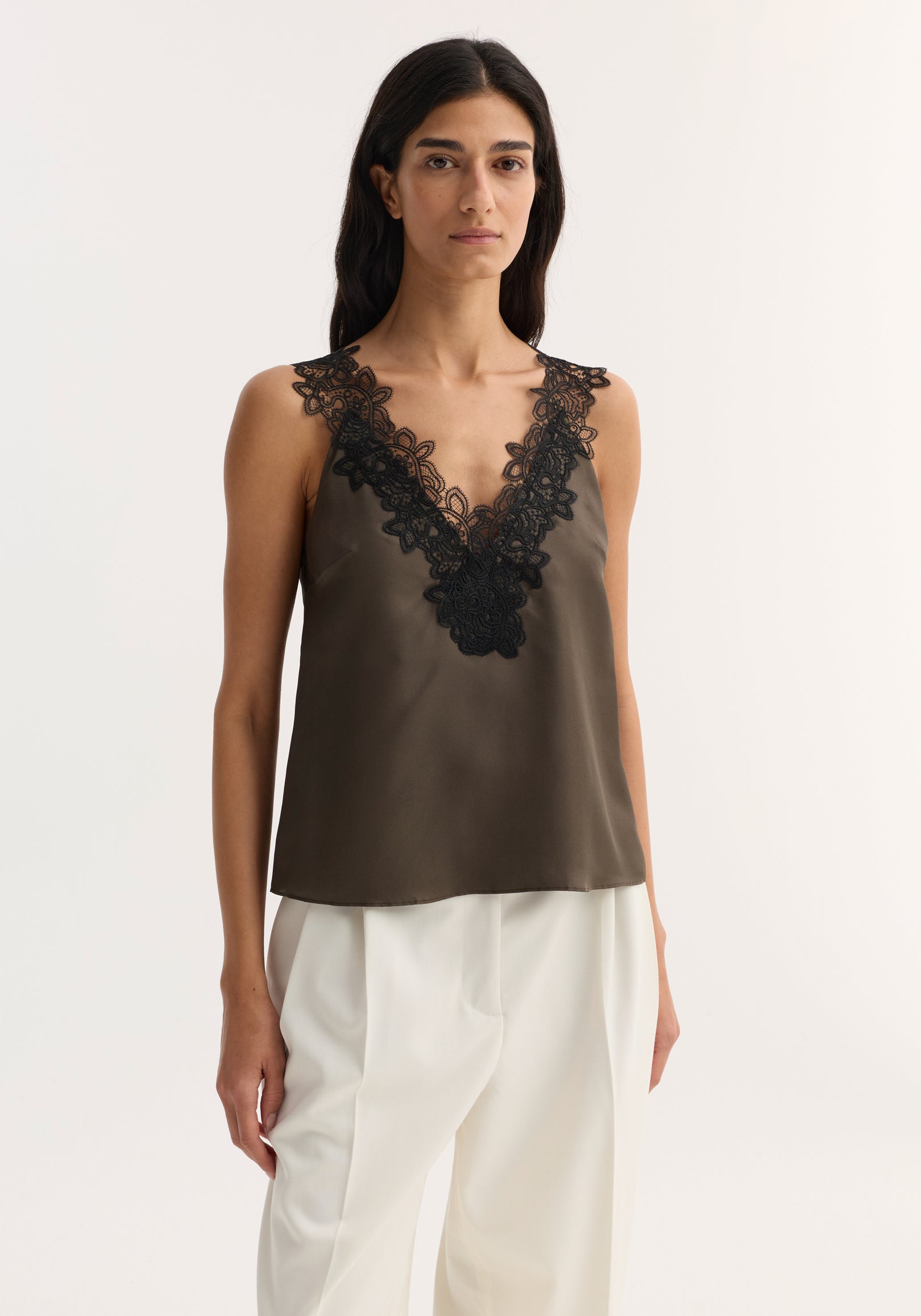 Woman wearing a sleeveless V-neck silk lace top | earth in a deep olive tone with intricate black lace detailing along the neckline and shoulders, paired with white trousers.