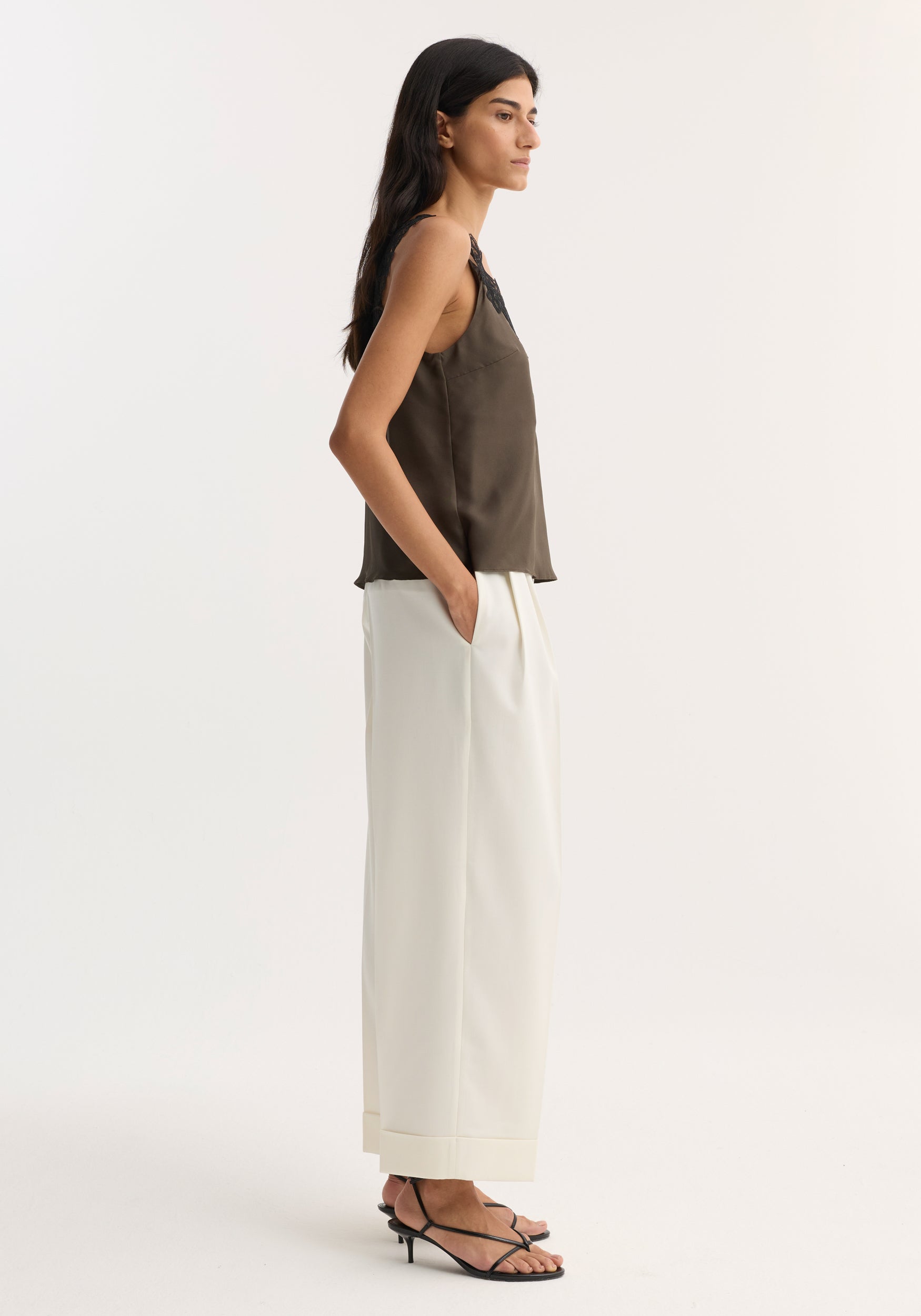 Side view of a woman wearing an earth-toned V-neck silk lace top paired with high-waisted white wide-leg pants and black strappy heels, styled with her hands in pockets.