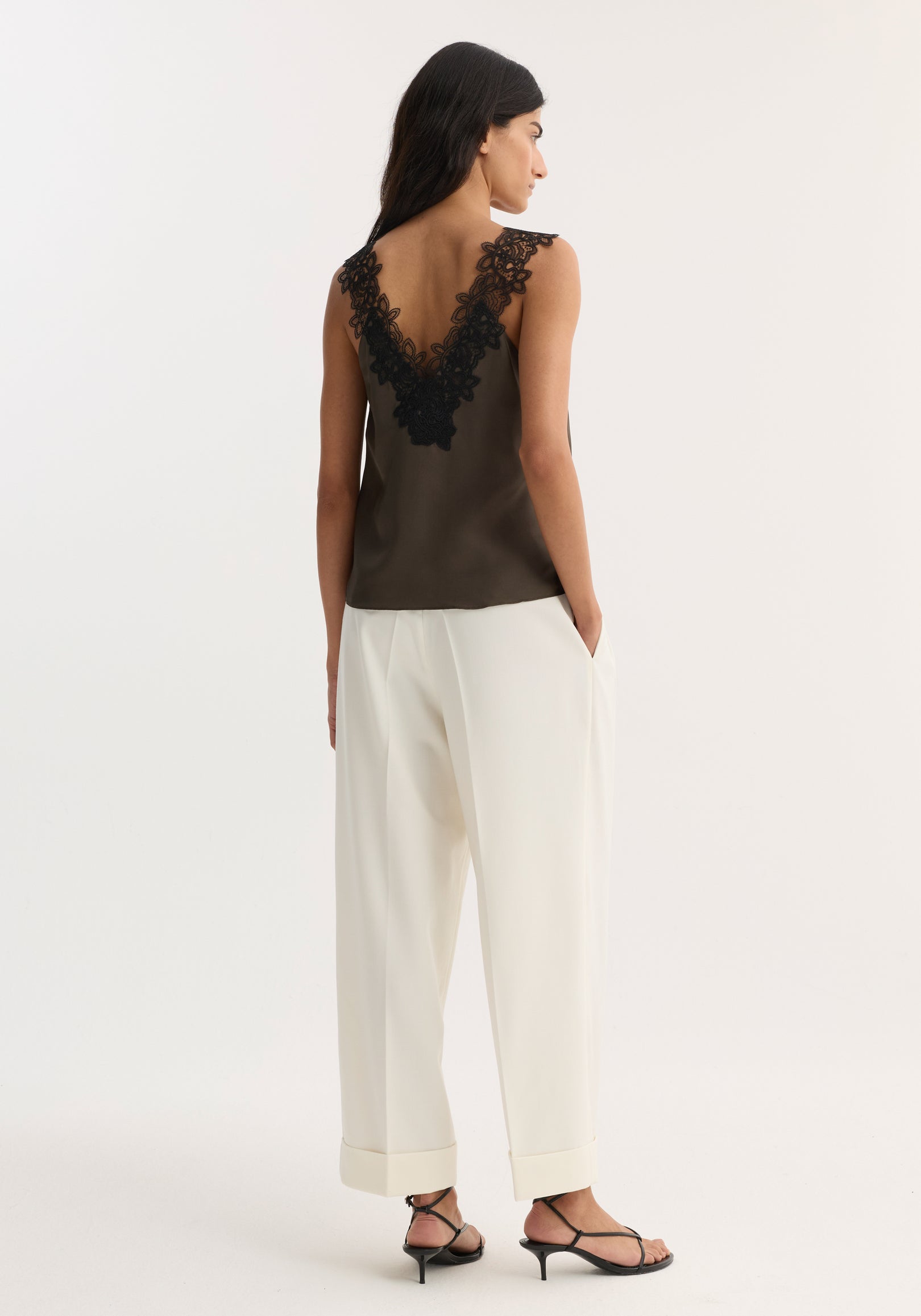 Woman wearing a V-neck silk lace top | earth in dark olive with intricate black lace detailing on the back, paired with loose white trousers and black heeled sandals, shown from behind.