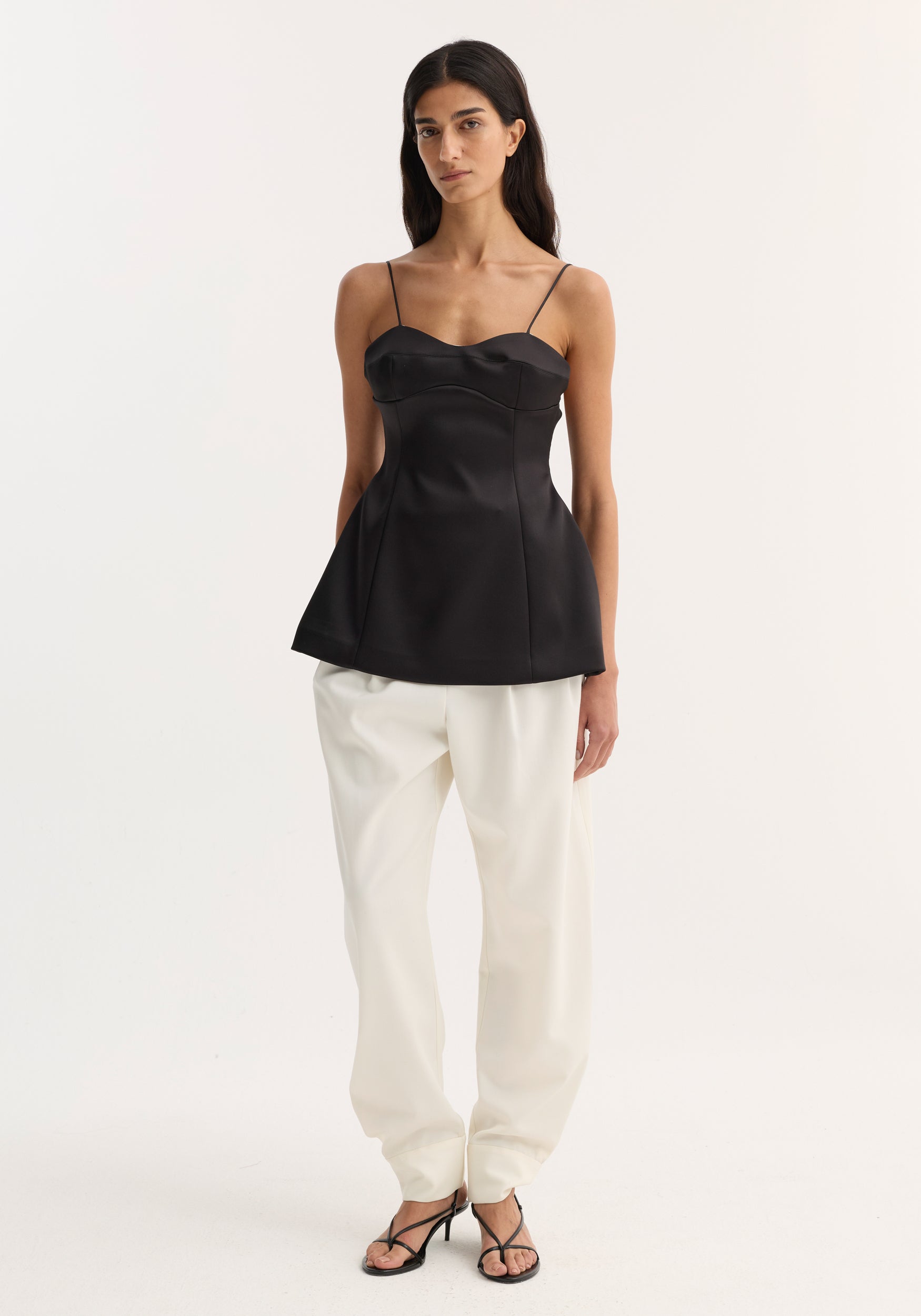 Woman wearing a fitted black bustier top | black with thin straps paired with loose white pants and black open-toe heels against a plain background.