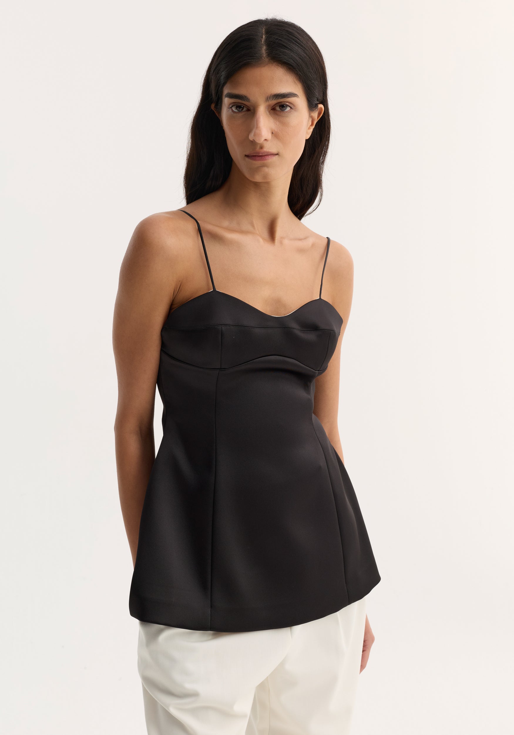 Woman wearing a black silky bustier top | black with thin spaghetti straps and a fitted bodice, paired with white pants against a plain light background.