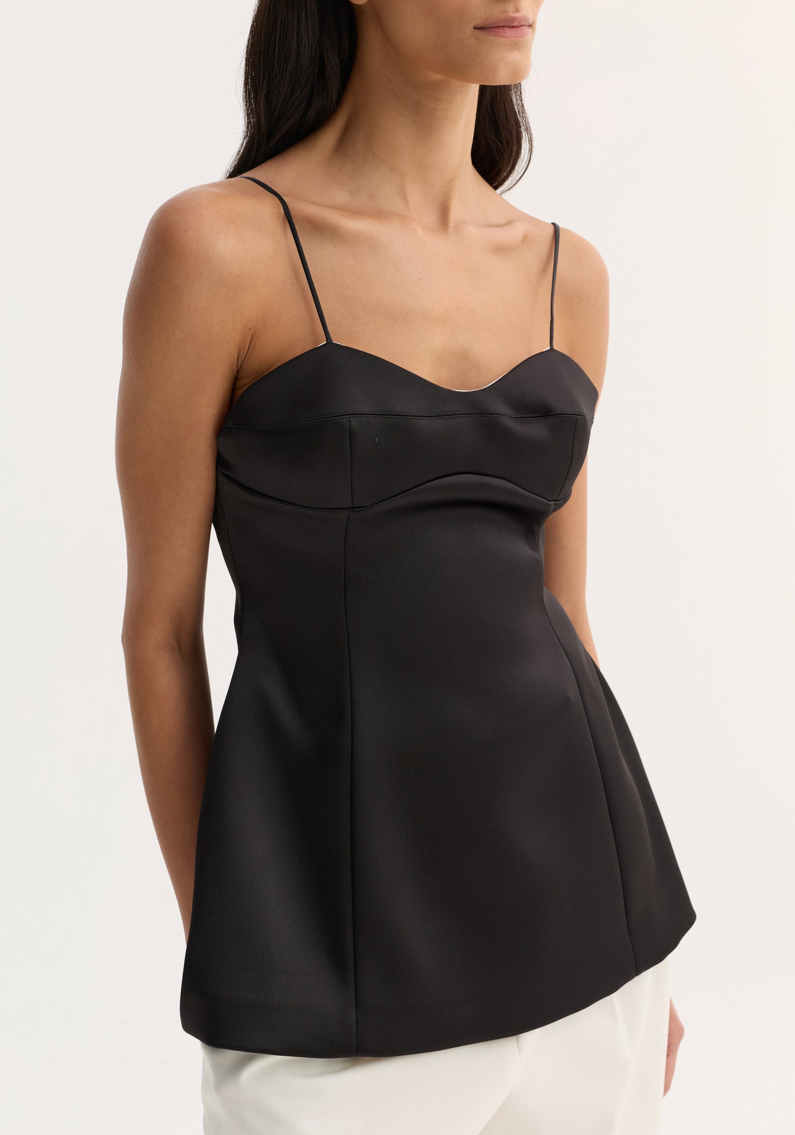 Woman wearing a sleek black bustier top | black with thin spaghetti straps and a fitted bodice, paired with white pants against a neutral background.