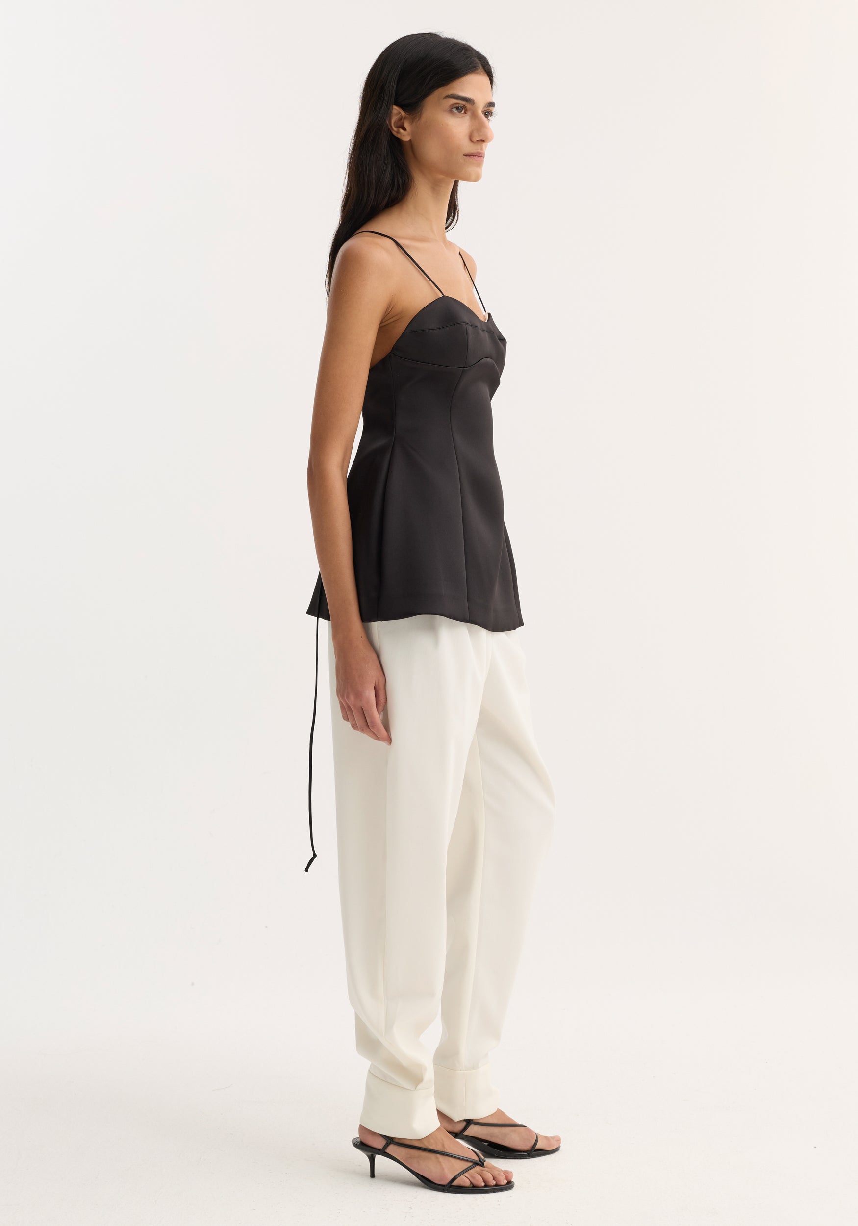 Side view of a woman wearing a fitted black Bustier top | black with thin straps, paired with loose white pants and black open-toe heels against a plain background.