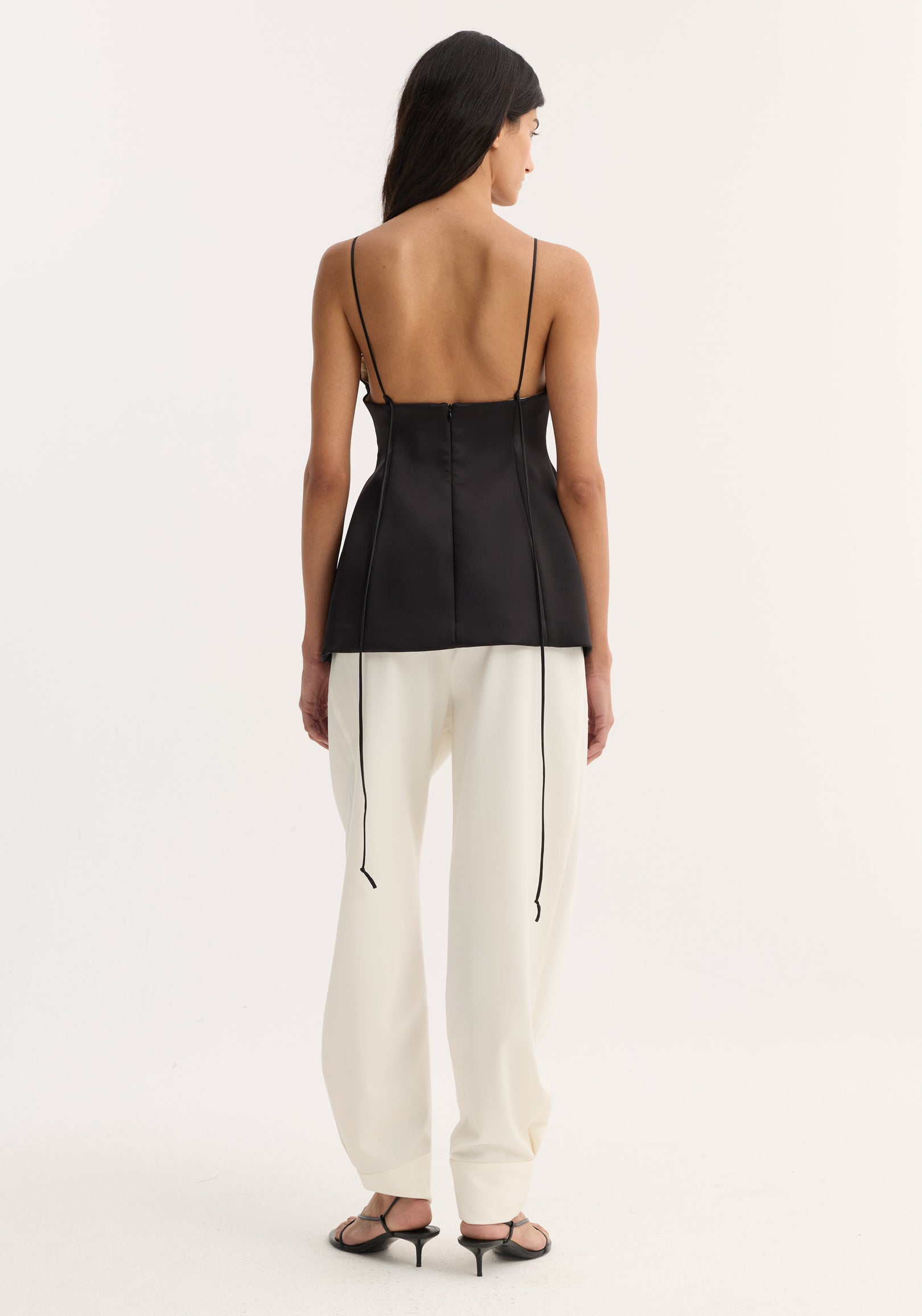 Woman wearing a black bustier top | black with thin straps and lace-up back detail, paired with white wide-leg pants and black heeled sandals, shown from behind.