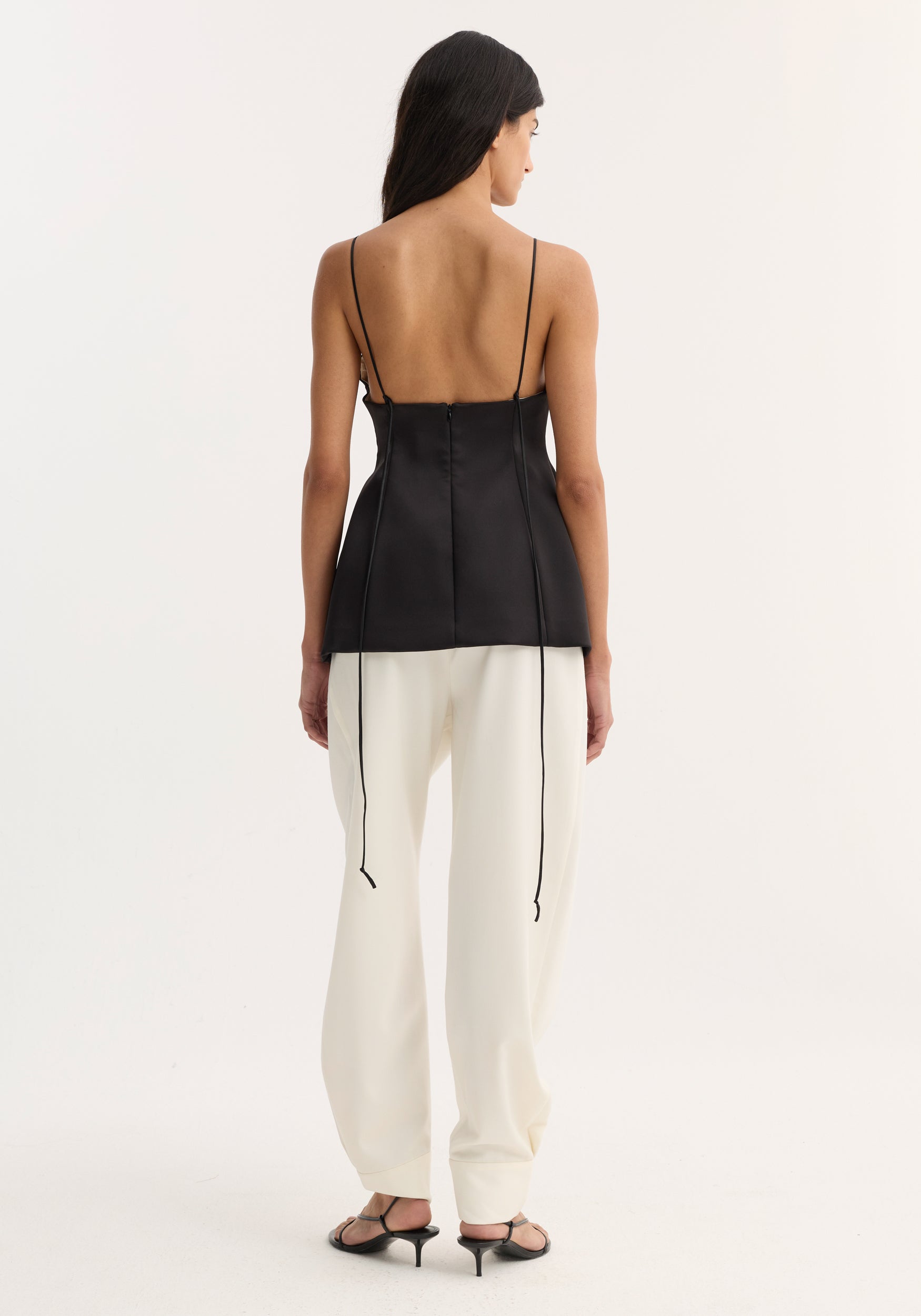 Woman wearing a black bustier top | black with thin straps and lace-up back detail, paired with white wide-leg pants and black heeled sandals, shown from behind.