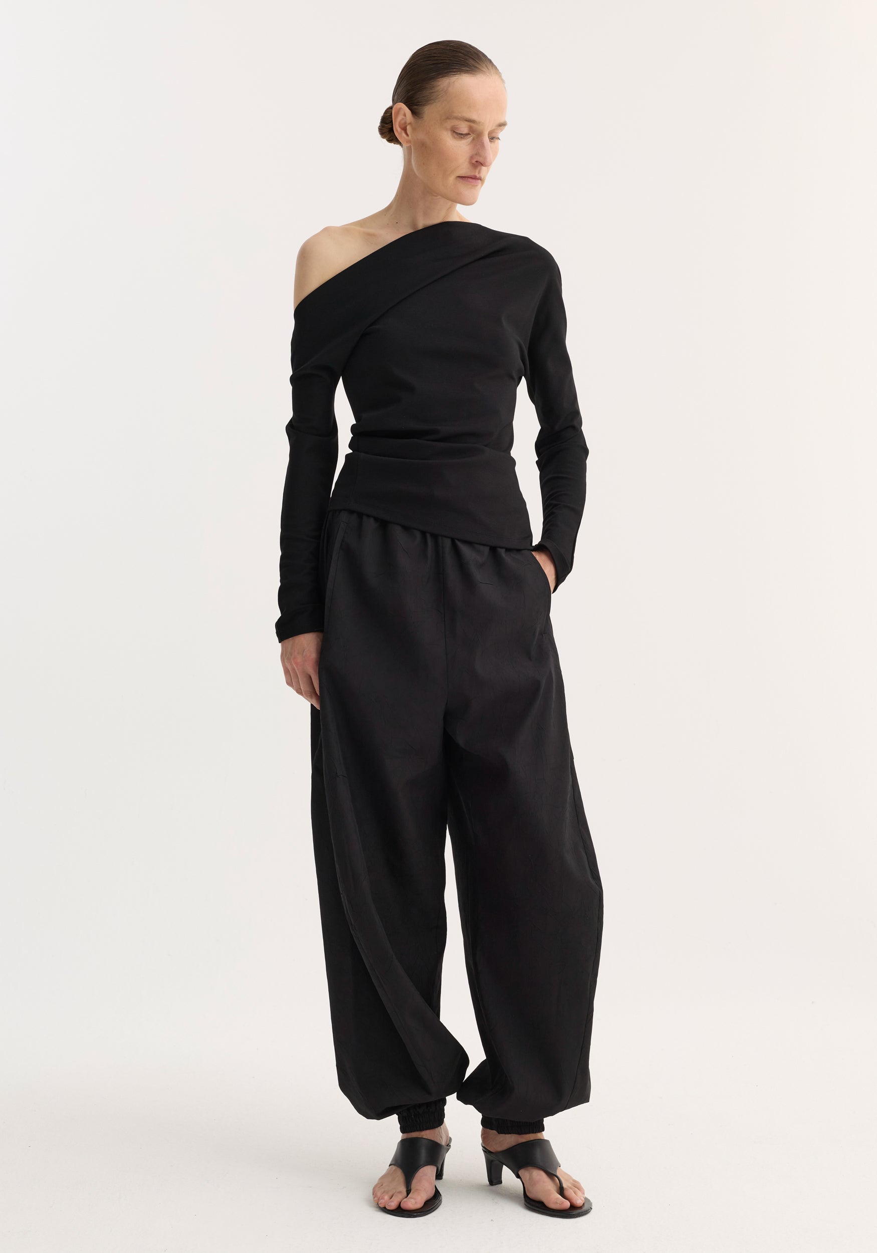 Woman wearing a long-sleeve twisted jersey top | black with an off-shoulder design paired with loose black pants and black open-toe heeled sandals against a plain background.