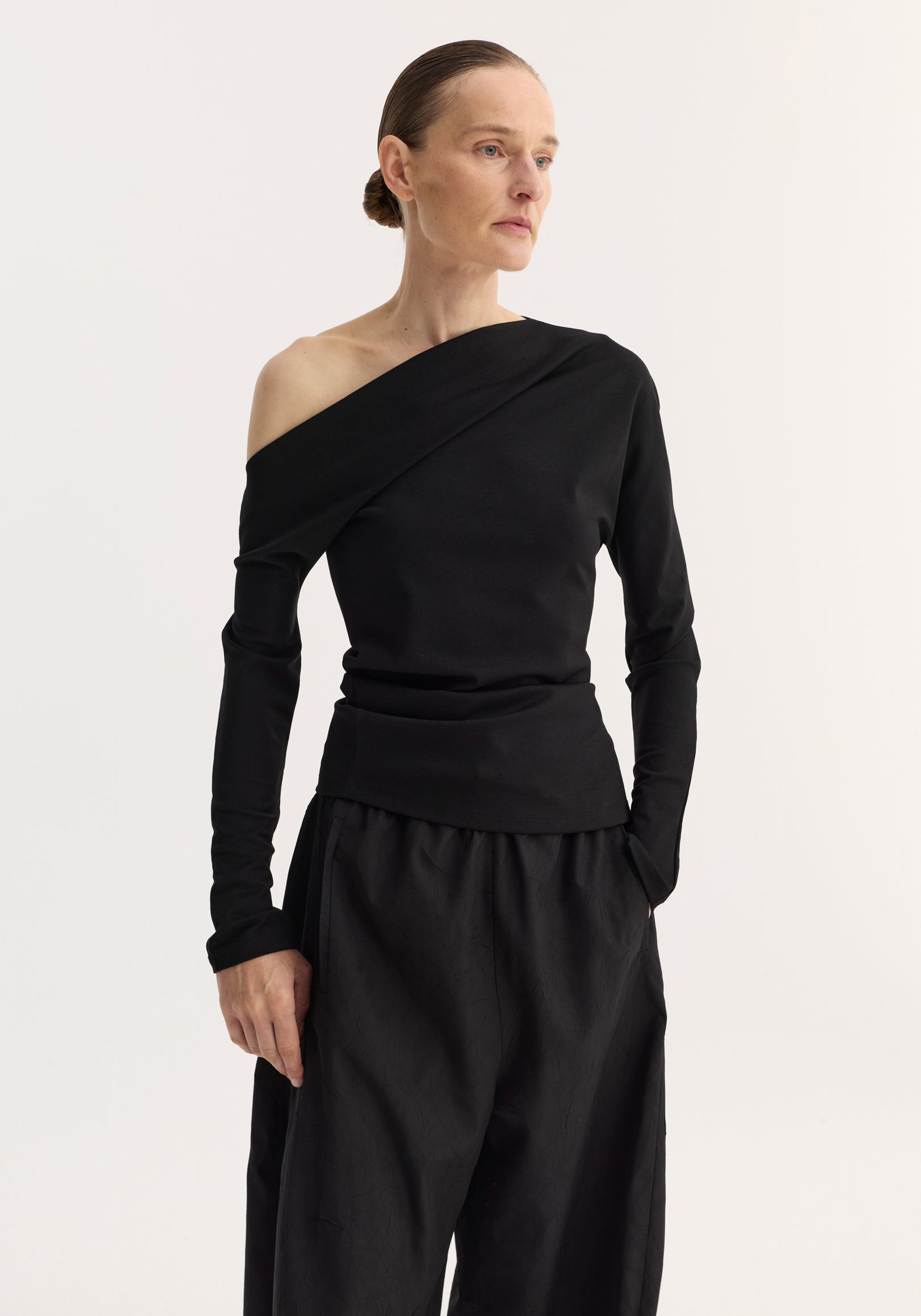 Woman wearing a black twisted jersey top | black with one off-shoulder long sleeve, paired with loose black pants, standing against a plain light background.