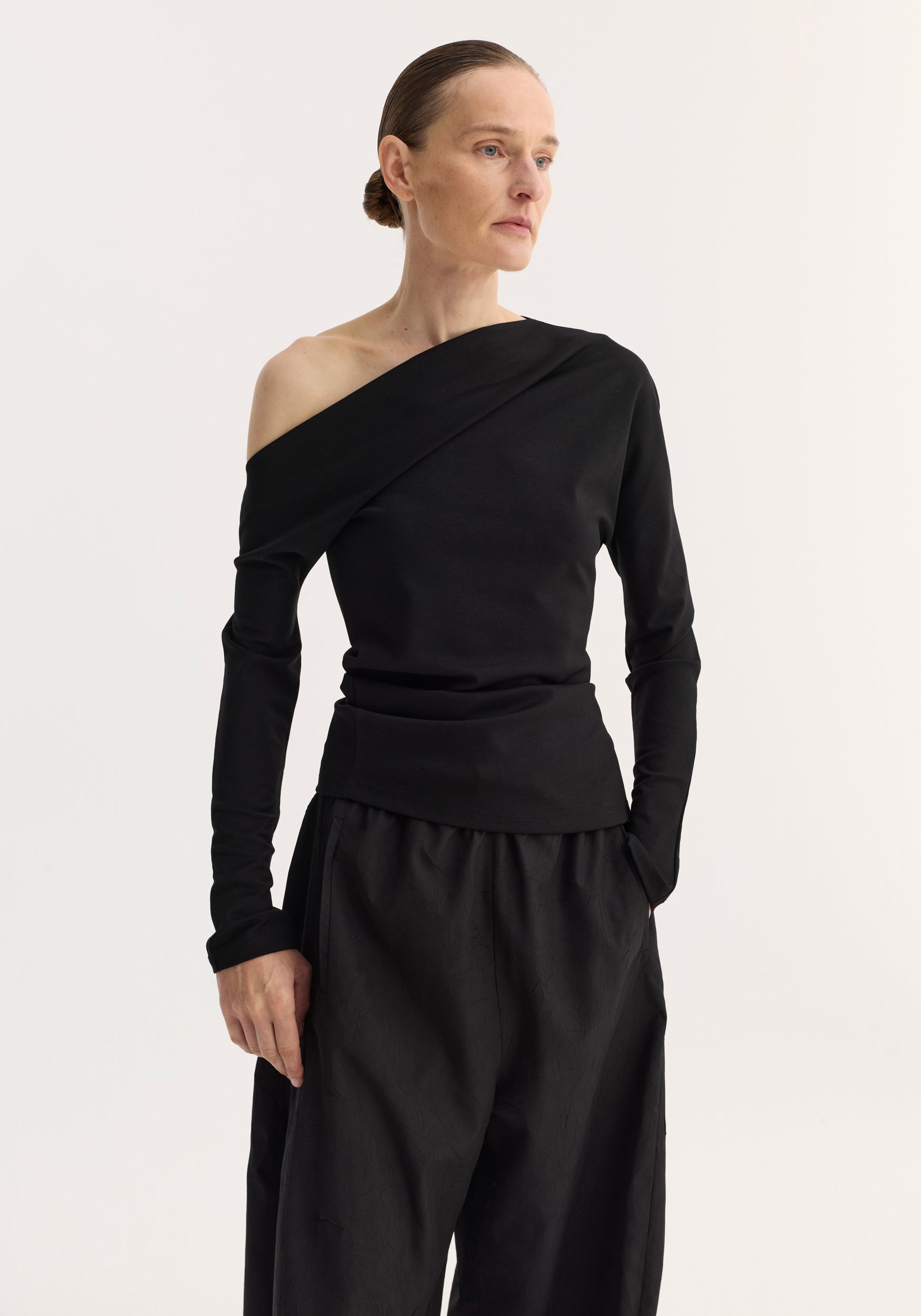 Woman wearing a black twisted jersey top | black with one off-shoulder long sleeve, paired with loose black pants, standing against a plain light background.