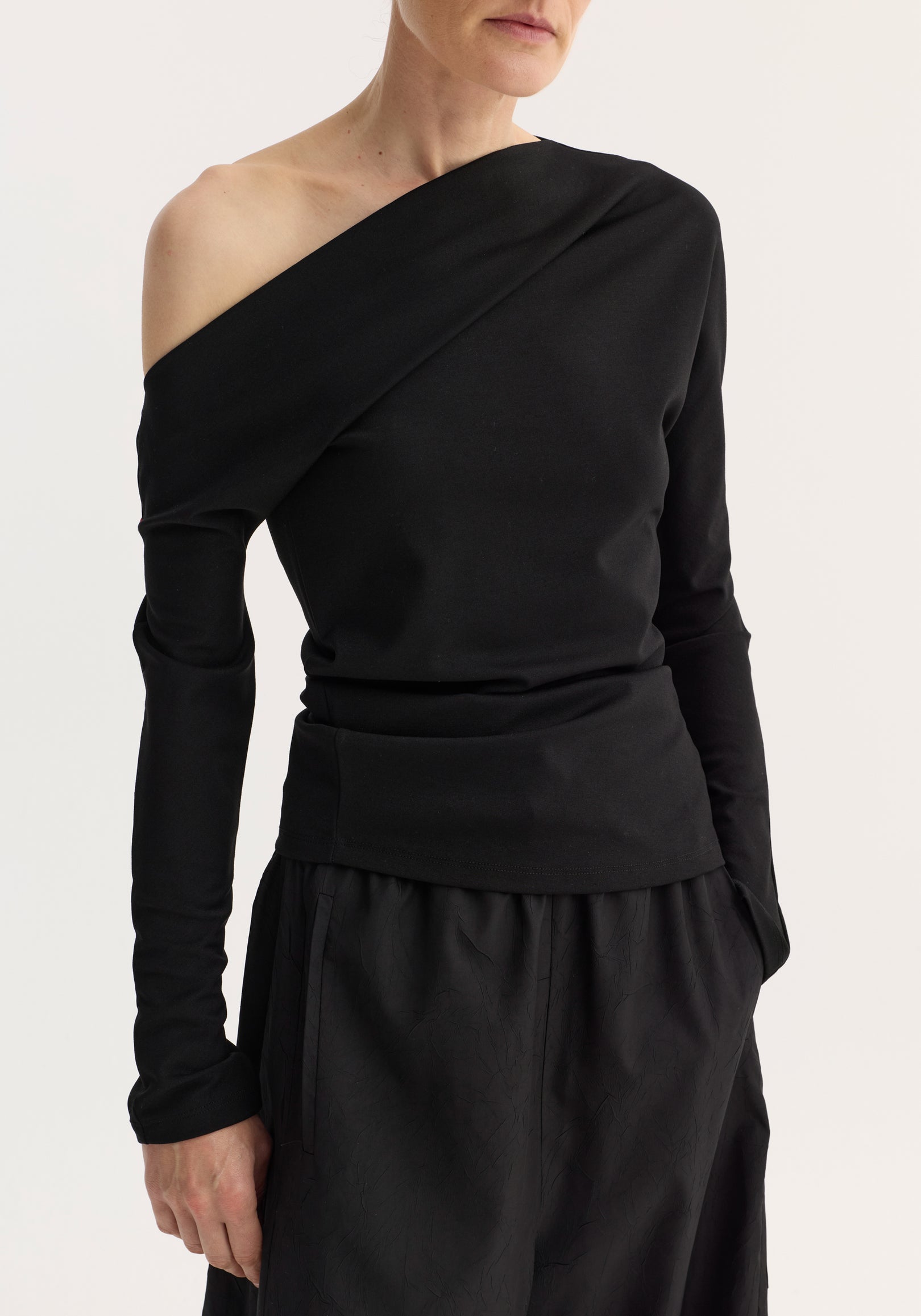 Woman wearing a black twisted jersey top | black with an asymmetrical off-shoulder neckline and long sleeves paired with black pants against a plain background.