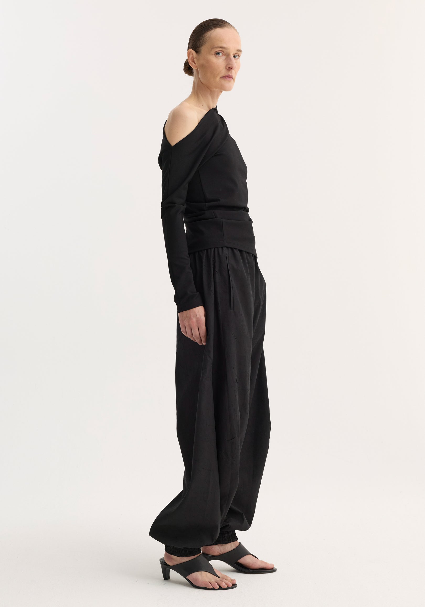 Model wearing a black twisted jersey top | black with long sleeves and a twisted off-shoulder neckline, paired with loose black pants and black heeled sandals against a plain white background.