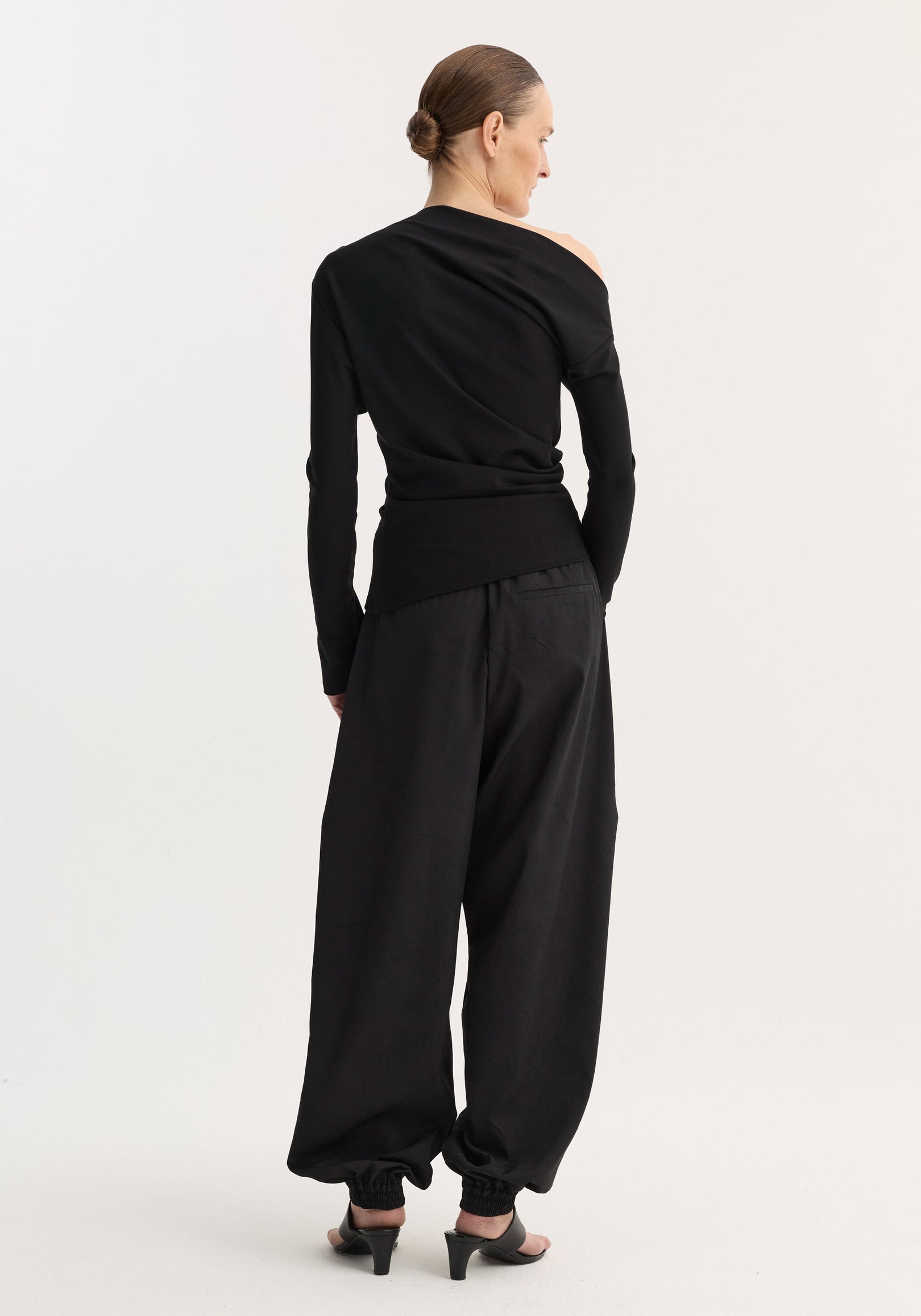 Woman standing facing away wearing a black twisted jersey top | black with an asymmetrical one-shoulder design, paired with loose black pants and black heeled sandals.