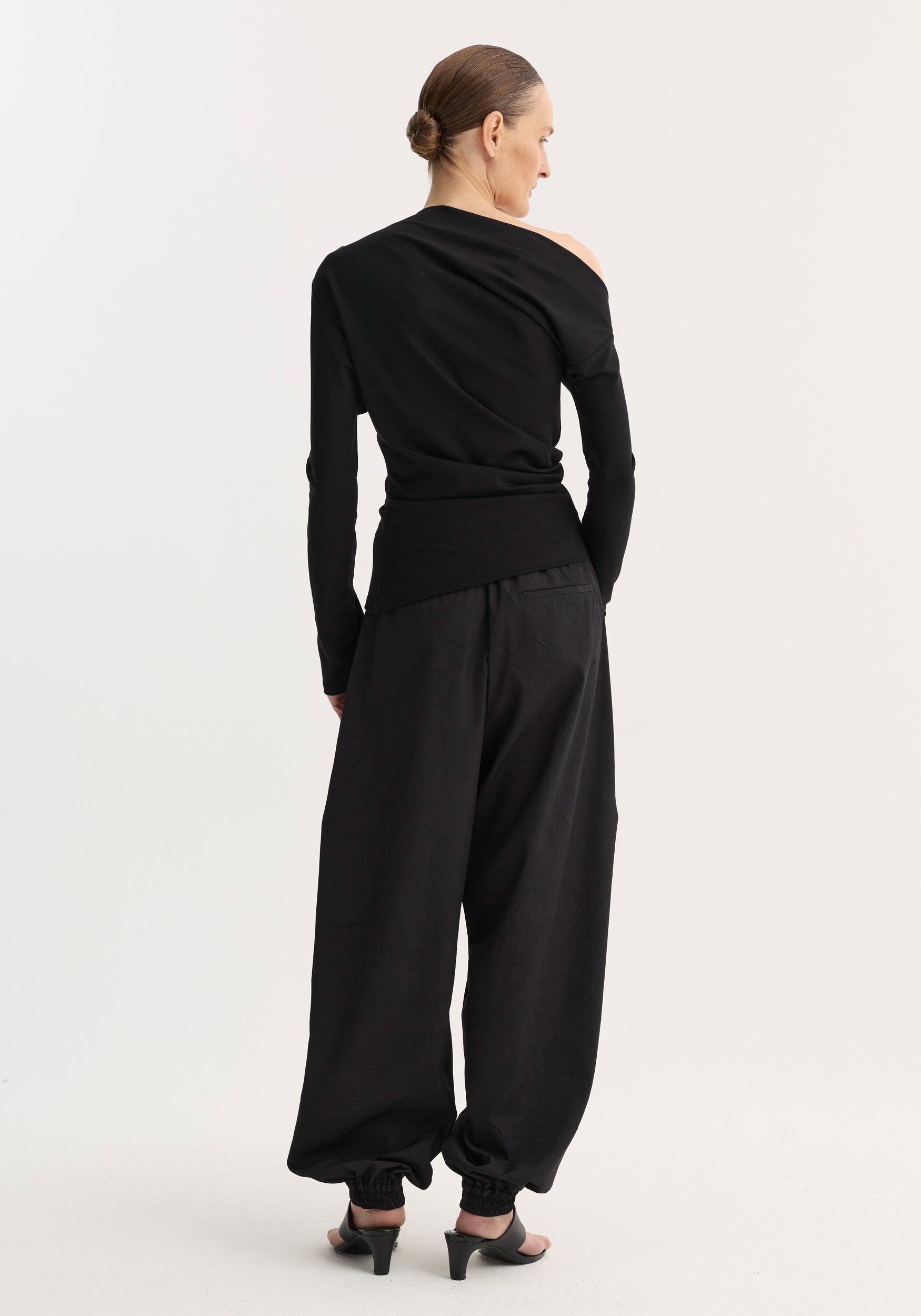 Woman standing facing away wearing a black twisted jersey top | black with an asymmetrical one-shoulder design, paired with loose black pants and black heeled sandals.