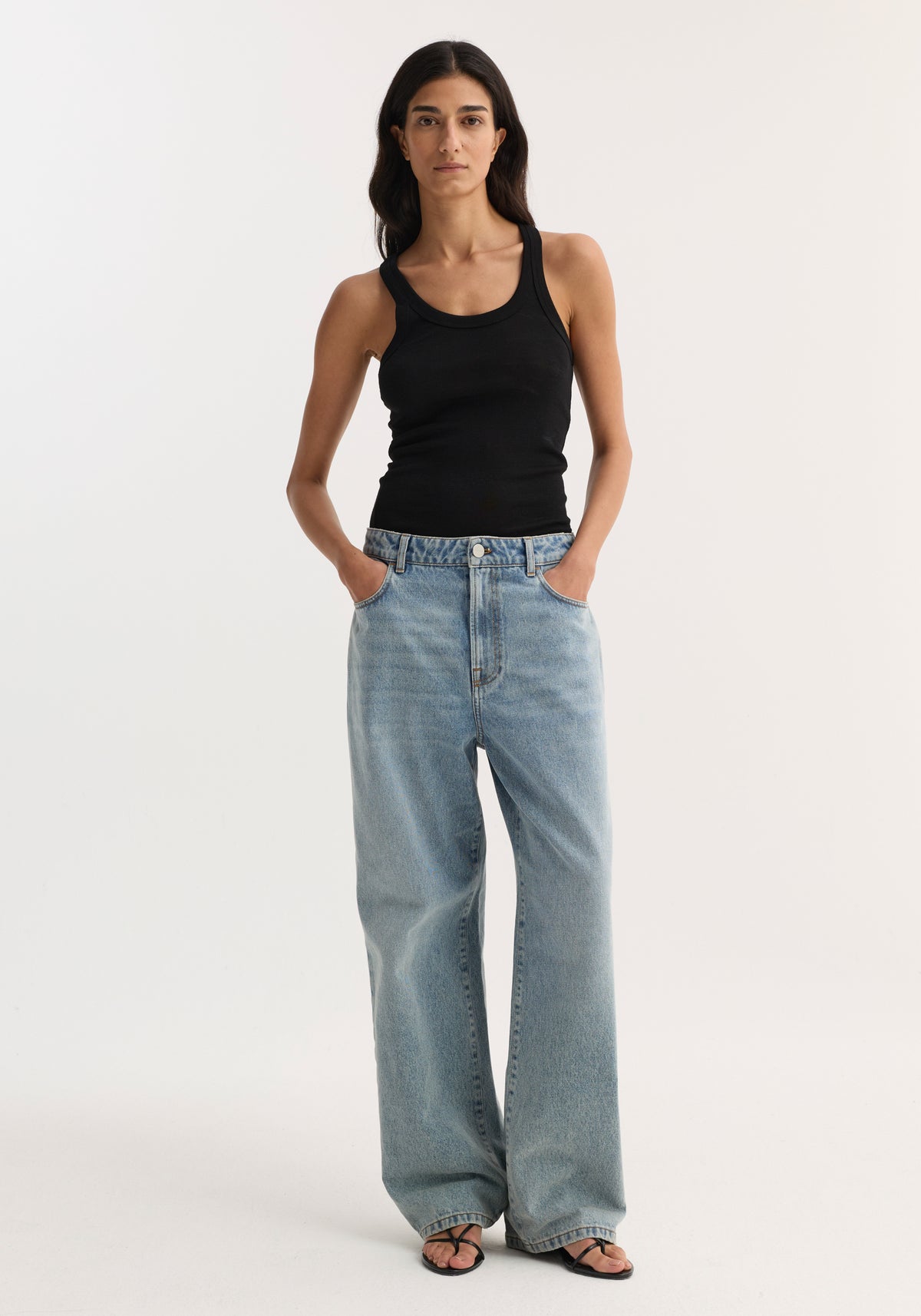 Woman wearing a black Merino scoop neck top | black paired with light blue wide-leg jeans and black sandals, standing with hands in pockets against a plain white background.
