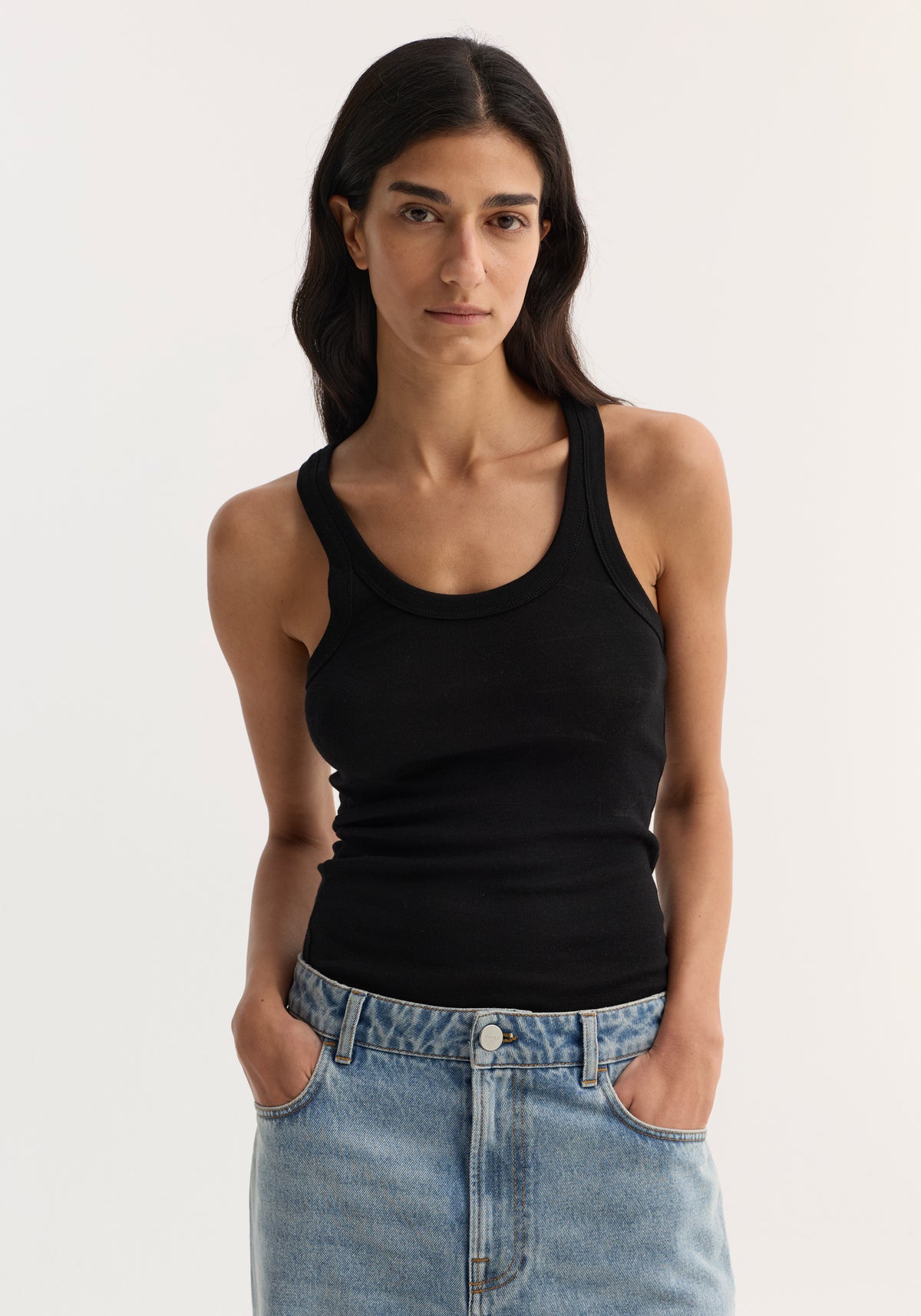 Woman wearing a fitted black Merino scoop neck top | black paired with light blue denim jeans, standing against a plain light background with hands in pockets.
