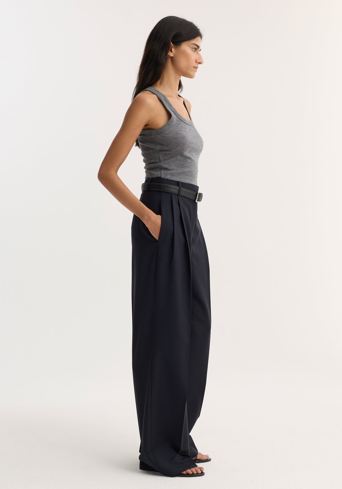 A woman wearing a fitted Merino scoop neck top | mid grey melange paired with high-waisted, wide-leg black trousers and a black belt, standing in profile against a plain background.