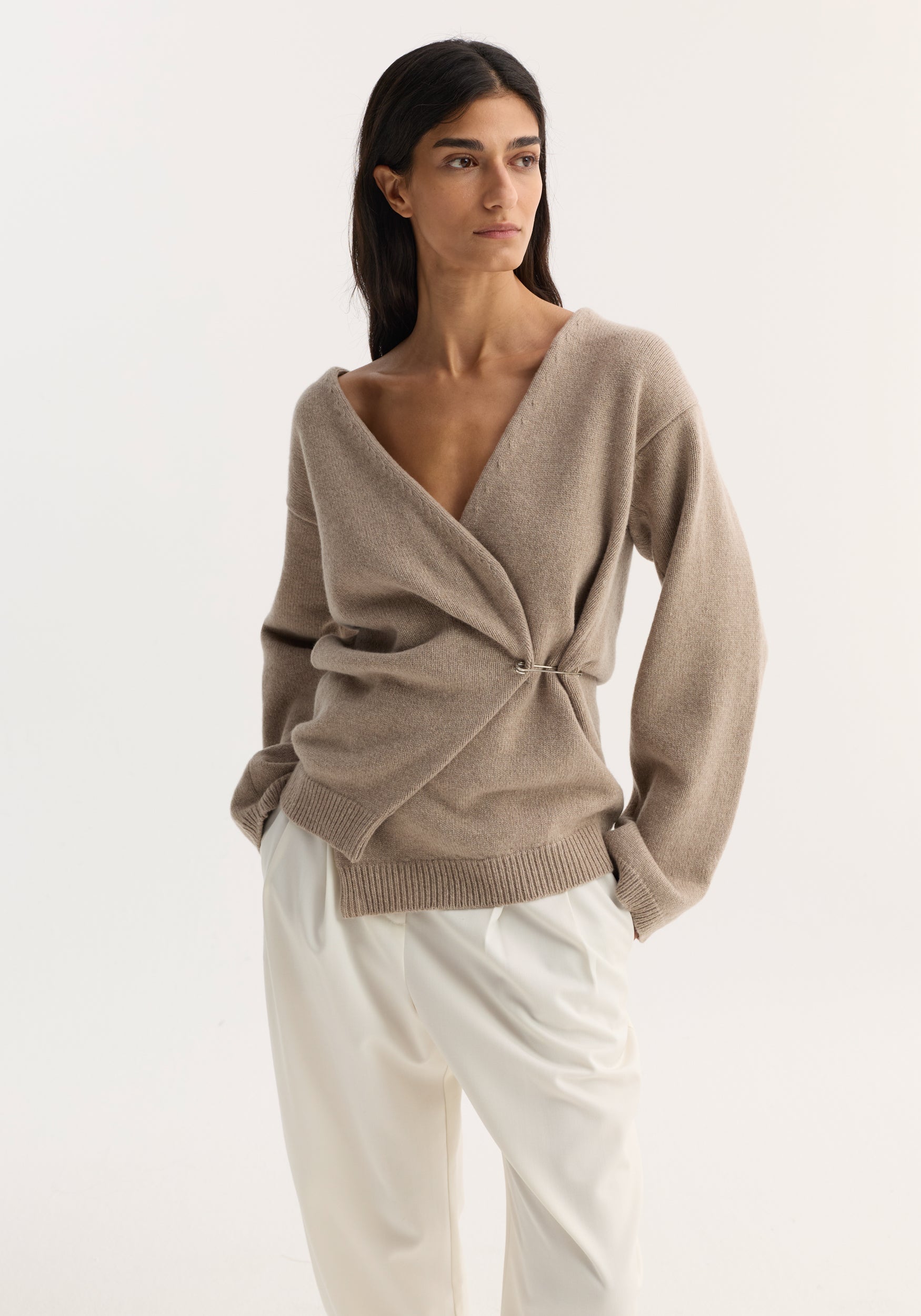 Woman wearing an off-shoulder wrap cardigan in light taupe with a relaxed fit and deep V-neck, paired with loose white pants against a plain background.