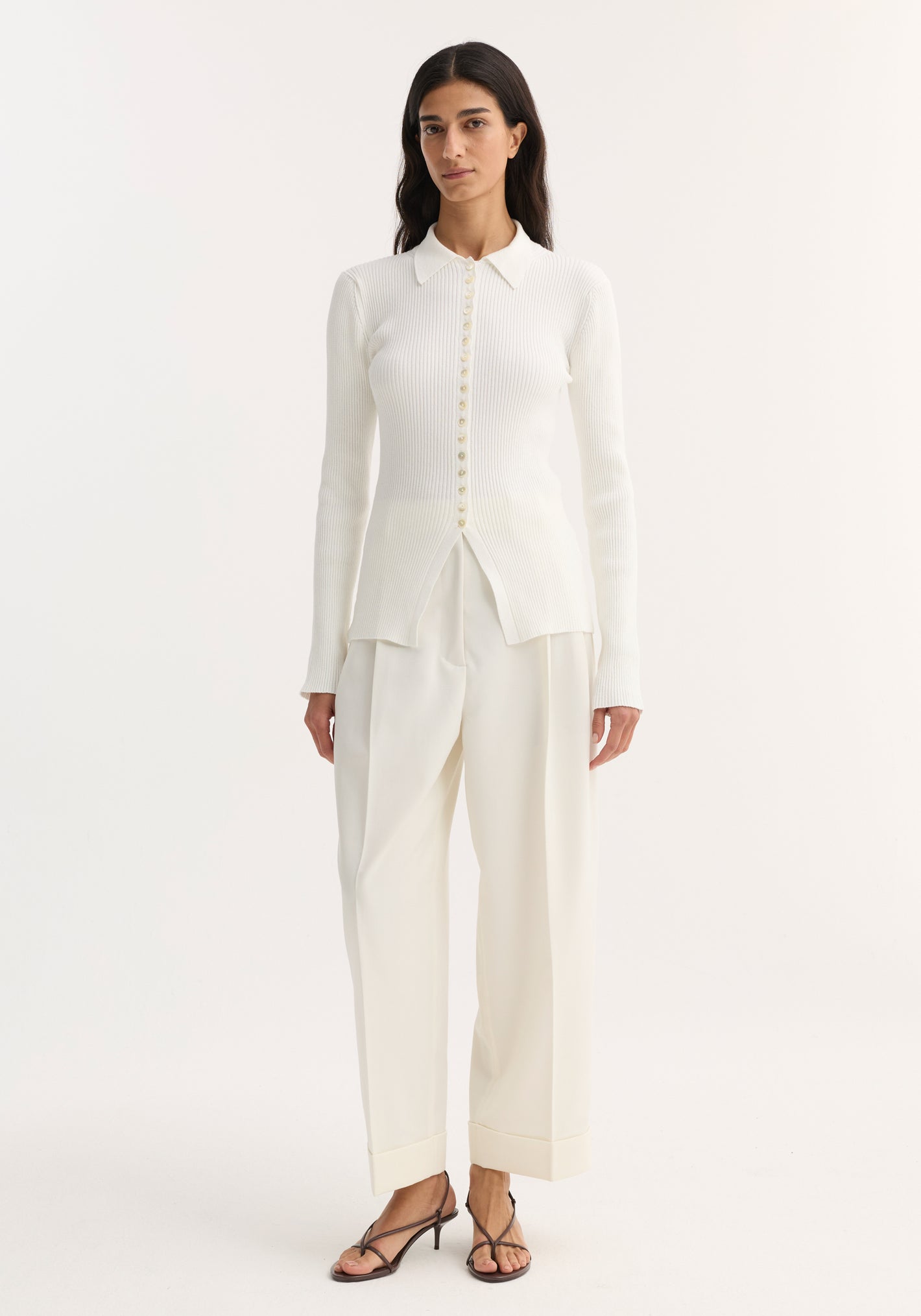 Model wears Elongated Rib Cardigan in off white, featuring a collar, ribbed texture, and front buttons ending at a hem slit, paired with high-waisted trousers.